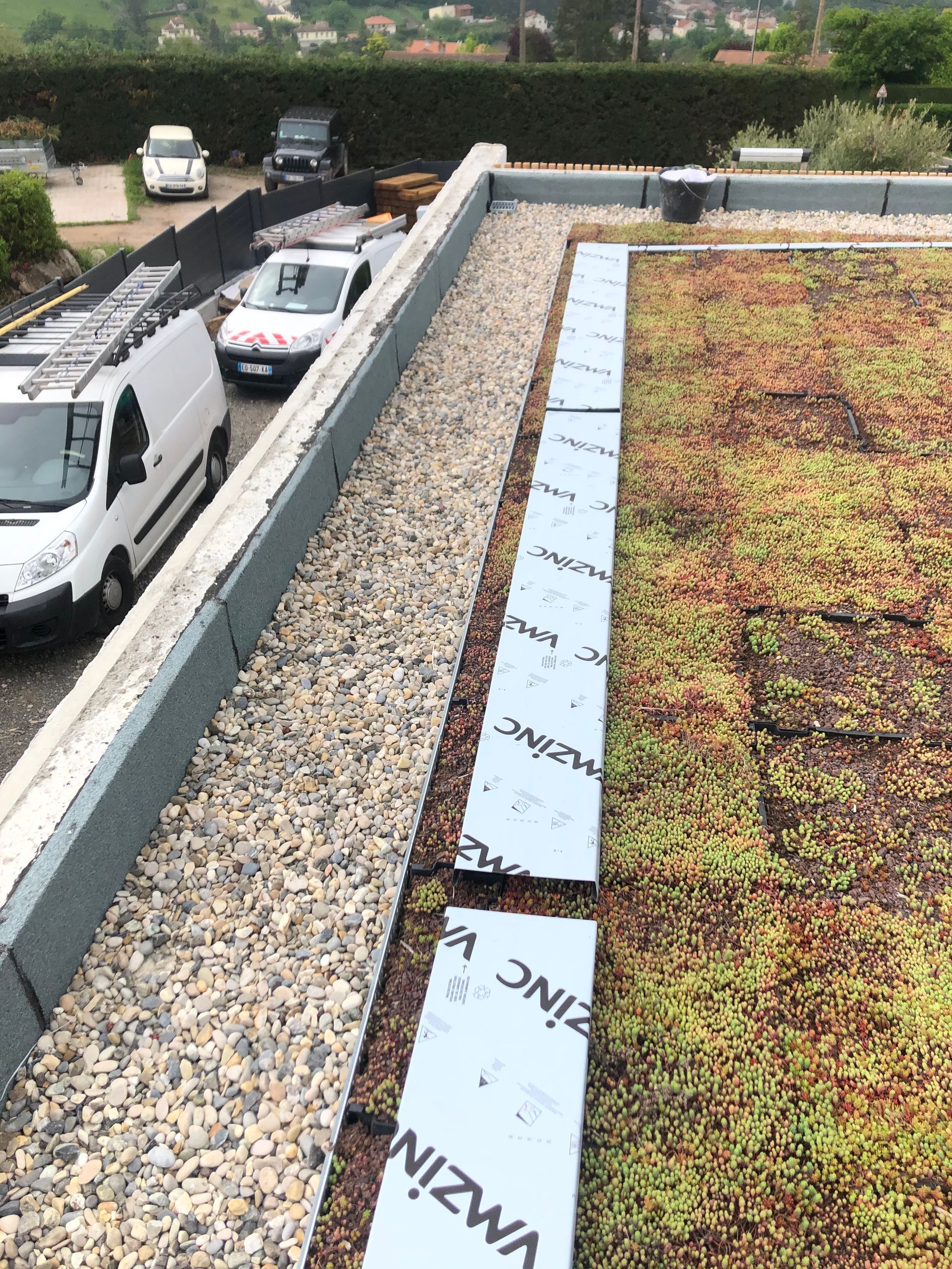 Toit-terrasse recouvert de gravier et de verdure, bordé de camionnettes garées et d'une route en contrebas.