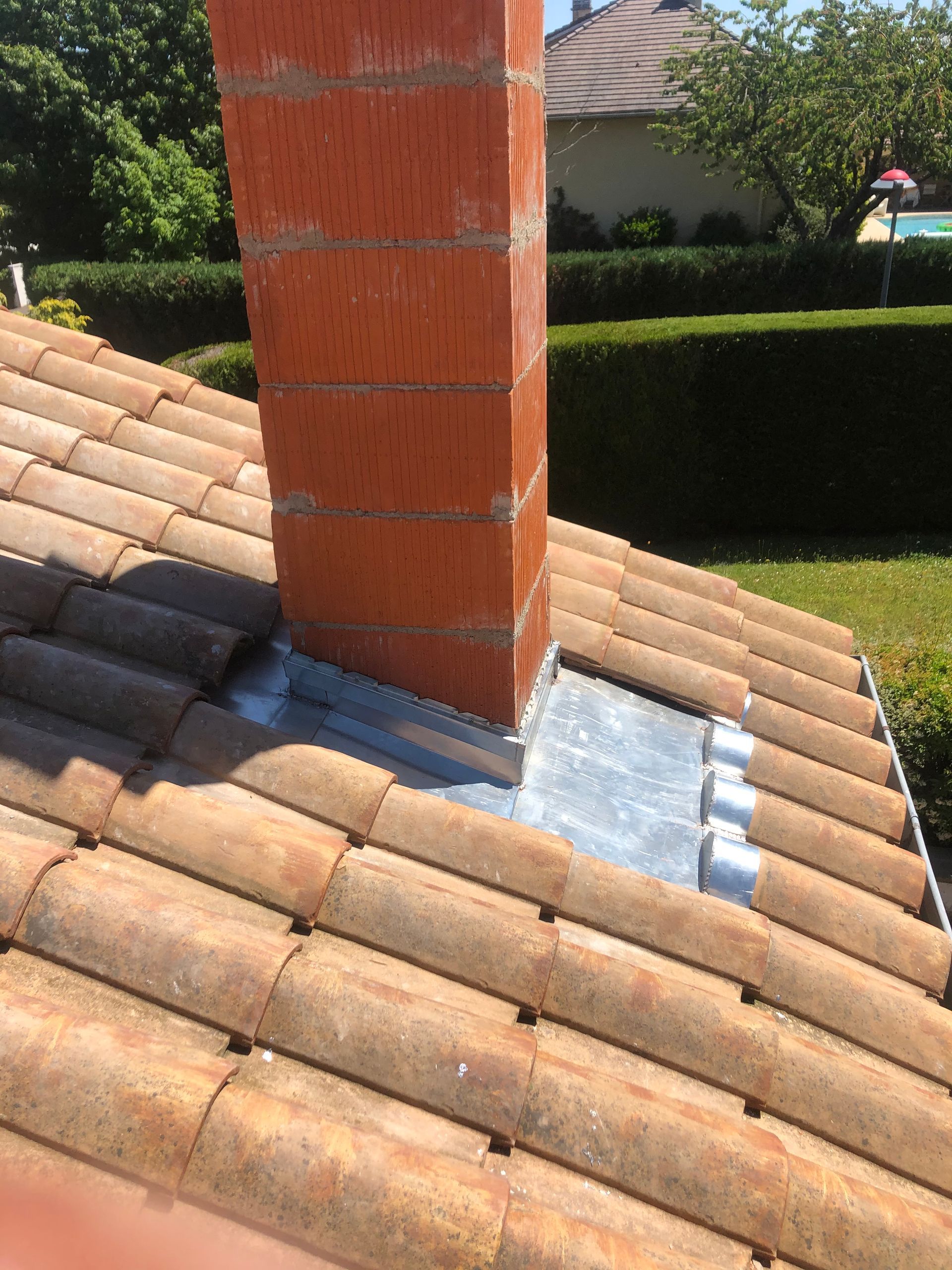 Toit de tuile avec cheminée en brique et solins métalliques argentés sous un soleil éclatant.