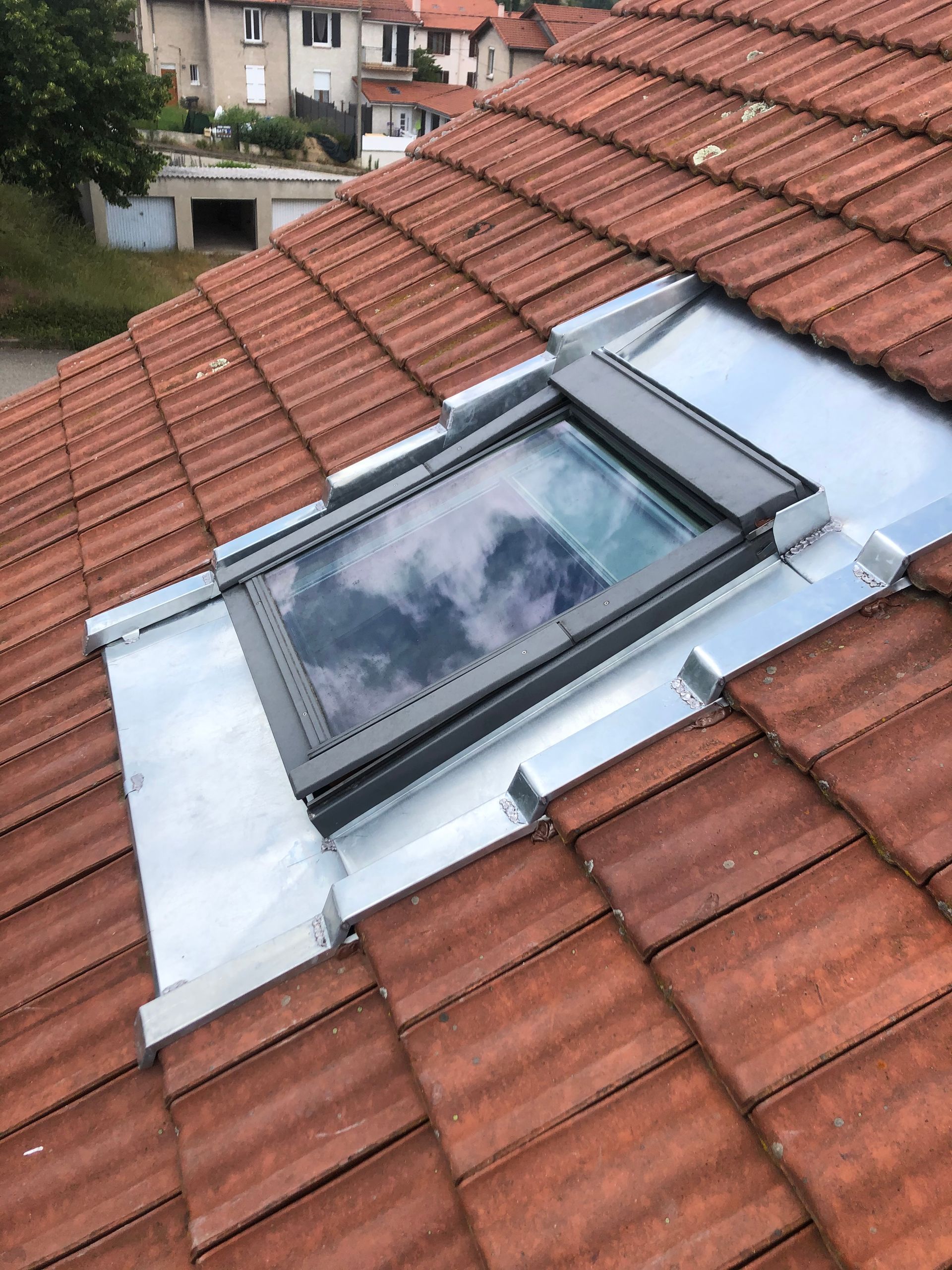 Fenêtre de toit dans une toiture de tuile rouge, entourée de tôles d'aluminium argentées.