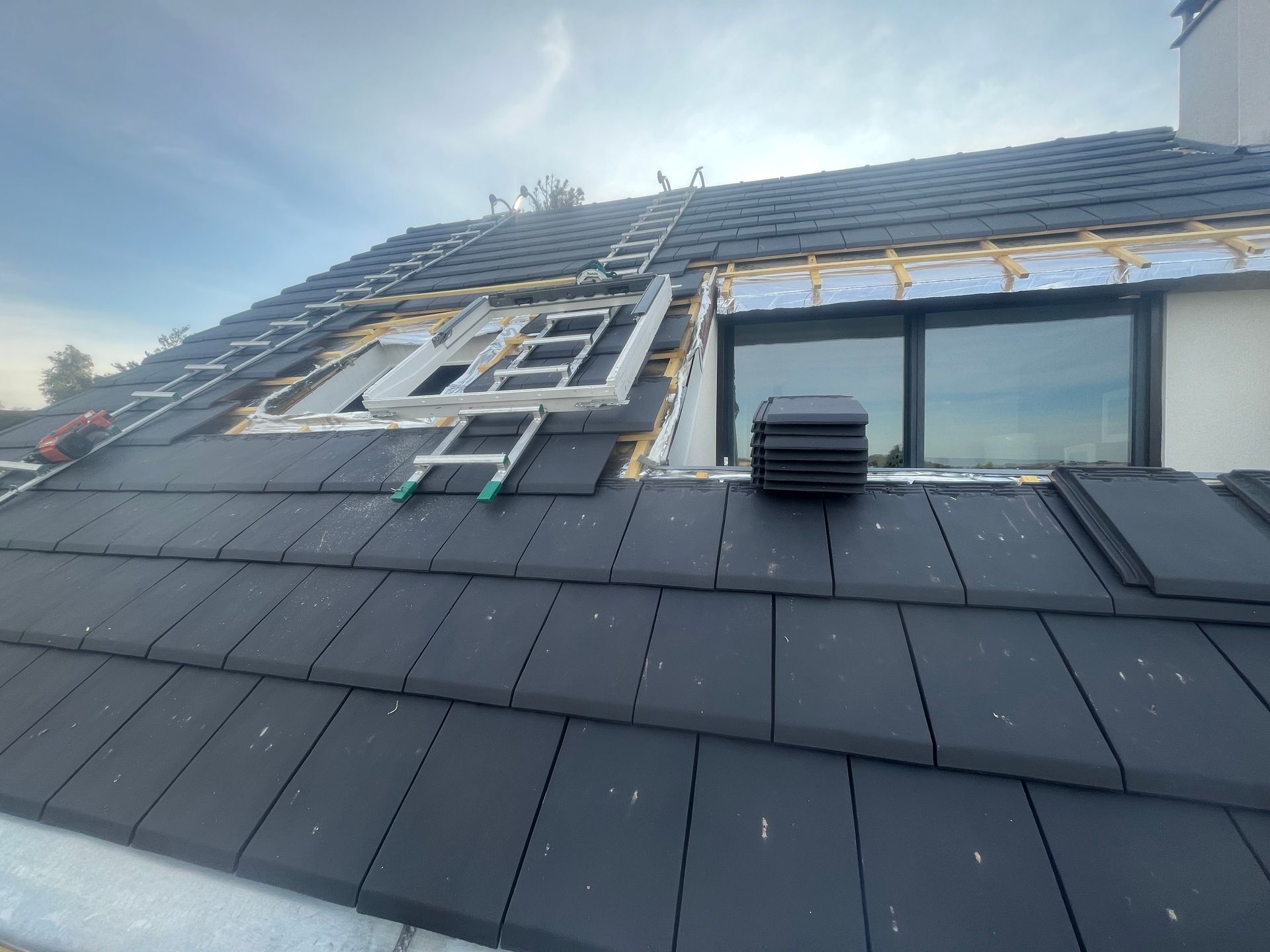 Toiture en réparation avec bardeaux noirs, sous-couche apparente, puits de lumière et cheminée d'évacuation sous un ciel bleu.