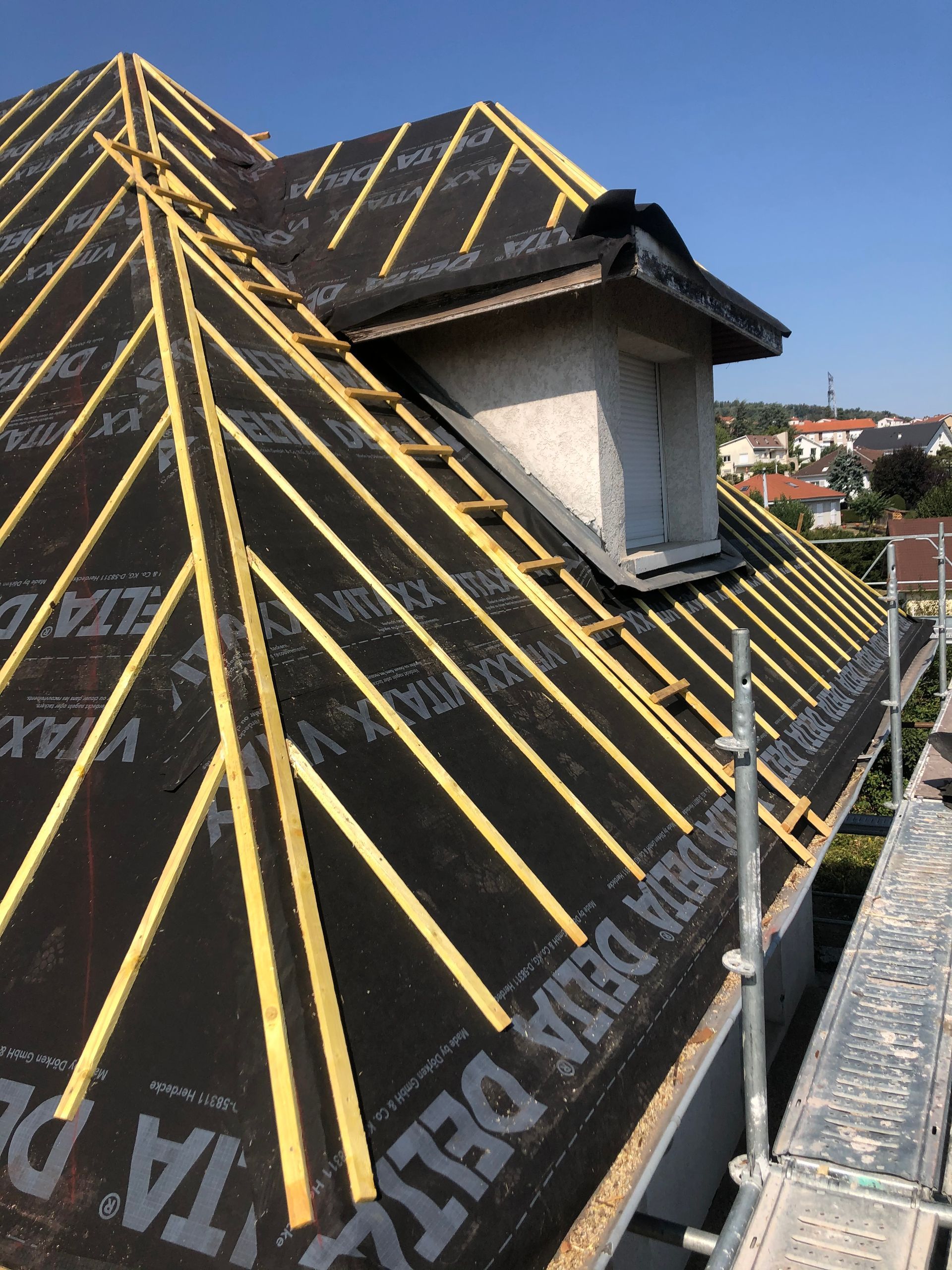 Toiture en construction avec étanchéité noire et liteaux jaunes à côté d'une lucarne.