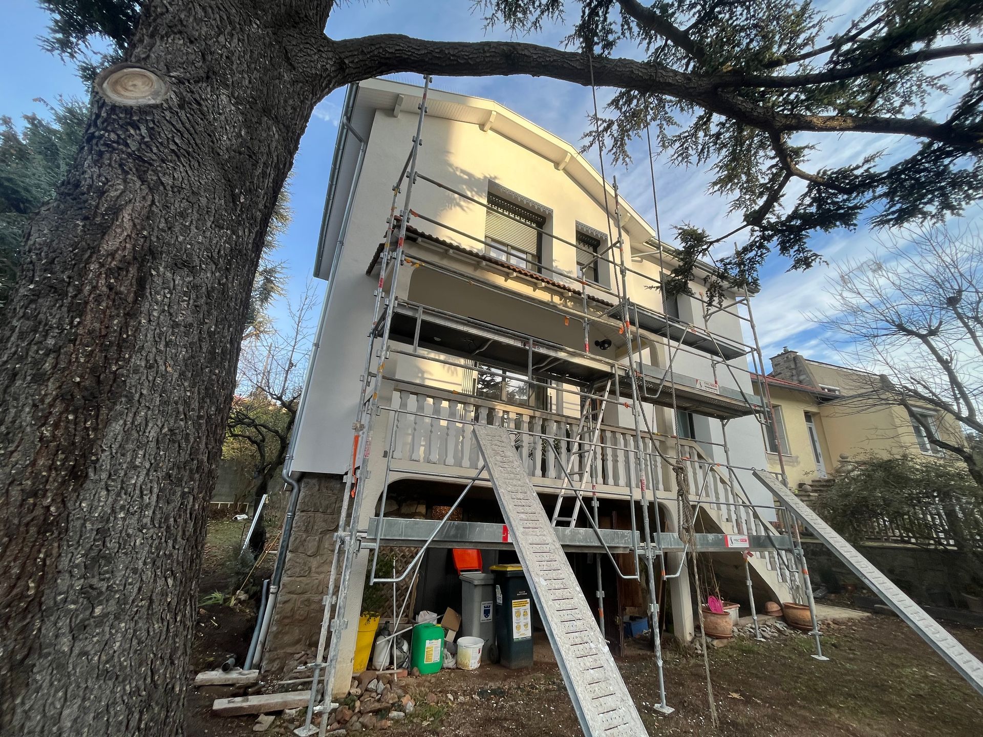 Maison en construction avec échafaudages et échelles à côté d'un grand arbre sous un ciel bleu.