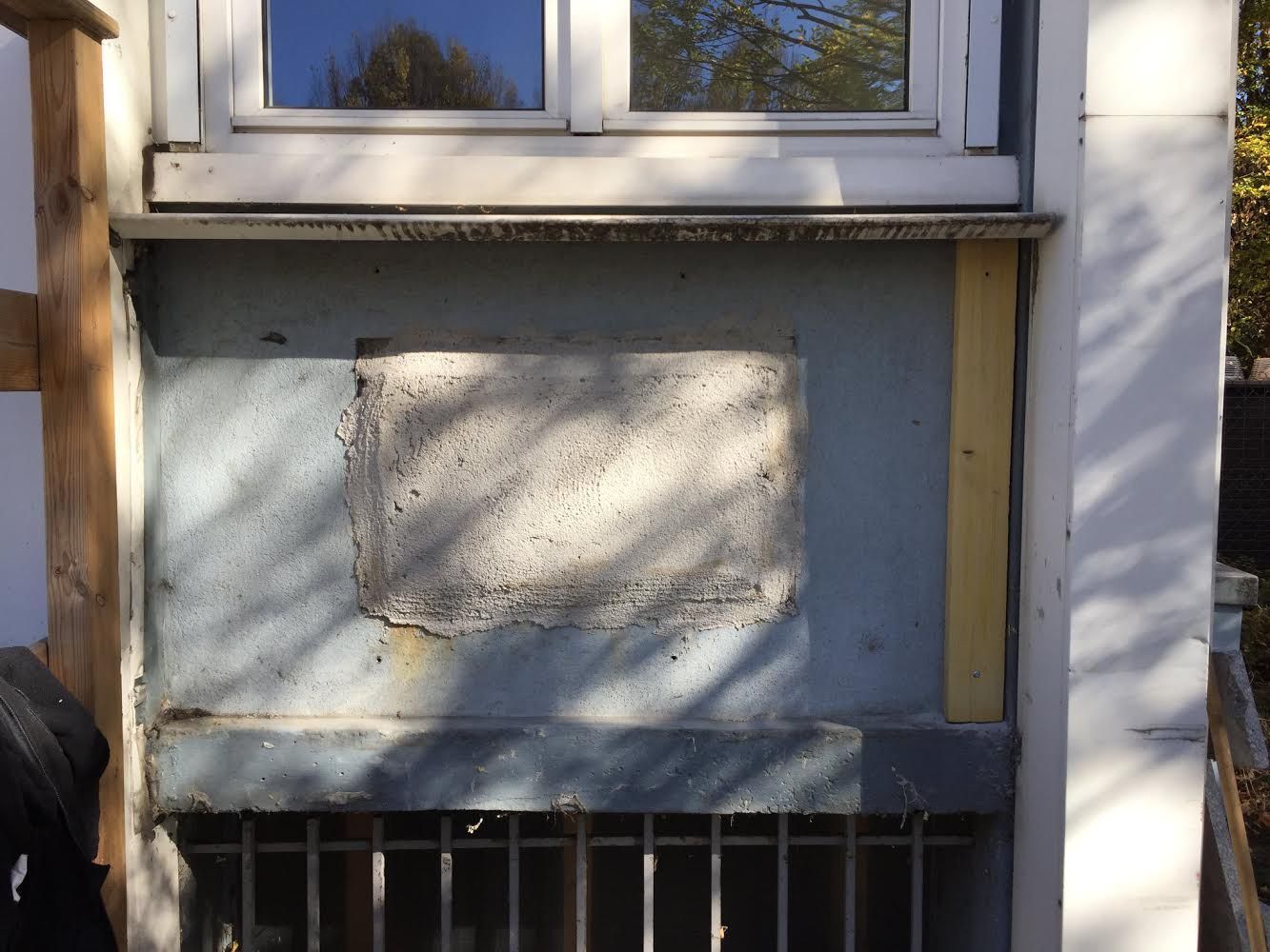 Mur d'une maison blanche avec une grande tache rectangulaire sous une fenêtre et une ouverture de cave avec des barreaux.