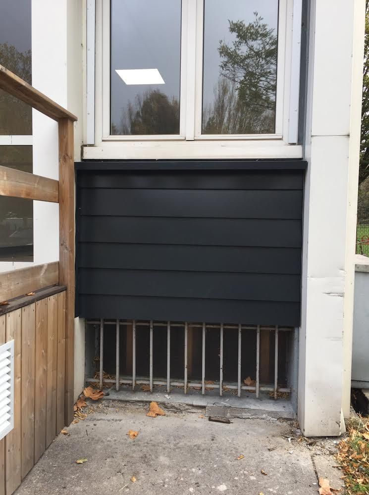 Fenêtre condamnée par des planches noires au-dessus d'une grille d'aération de sous-sol, avec une rambarde et une clôture en bois à côté.