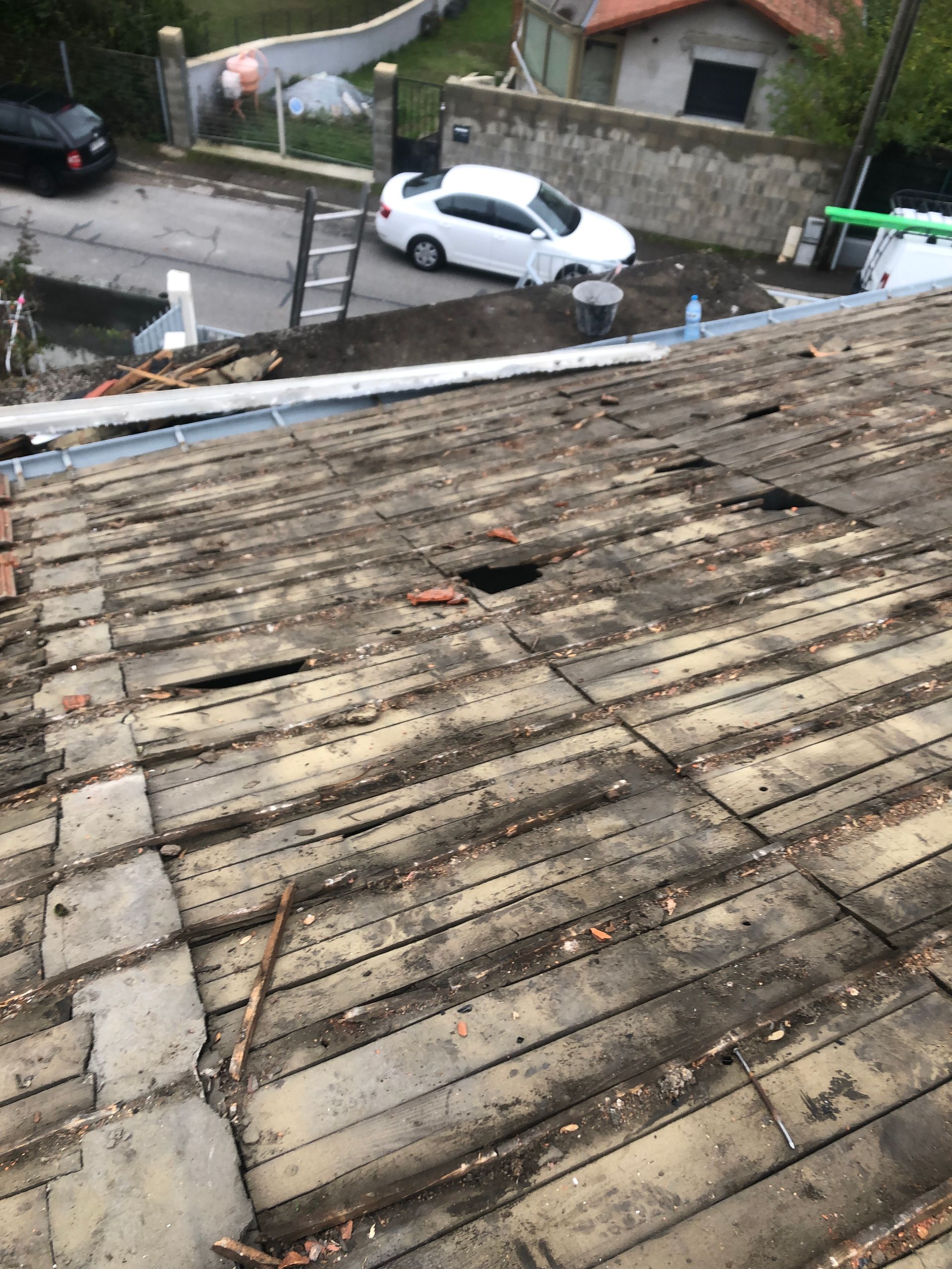 Toit de tuiles usées par le temps, avec une petite partie endommagée, donnant sur une rue avec une voiture blanche et un mur de pierre.