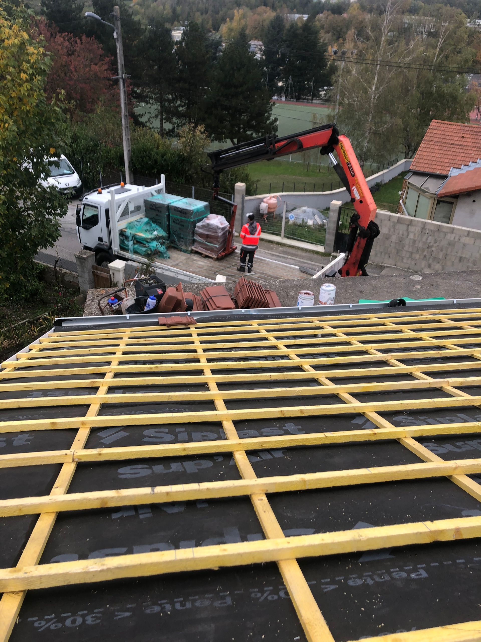 Construction d'un toit avec une grue orange, des ouvriers et des matériaux empilés à côté d'une maison dans une zone boisée.