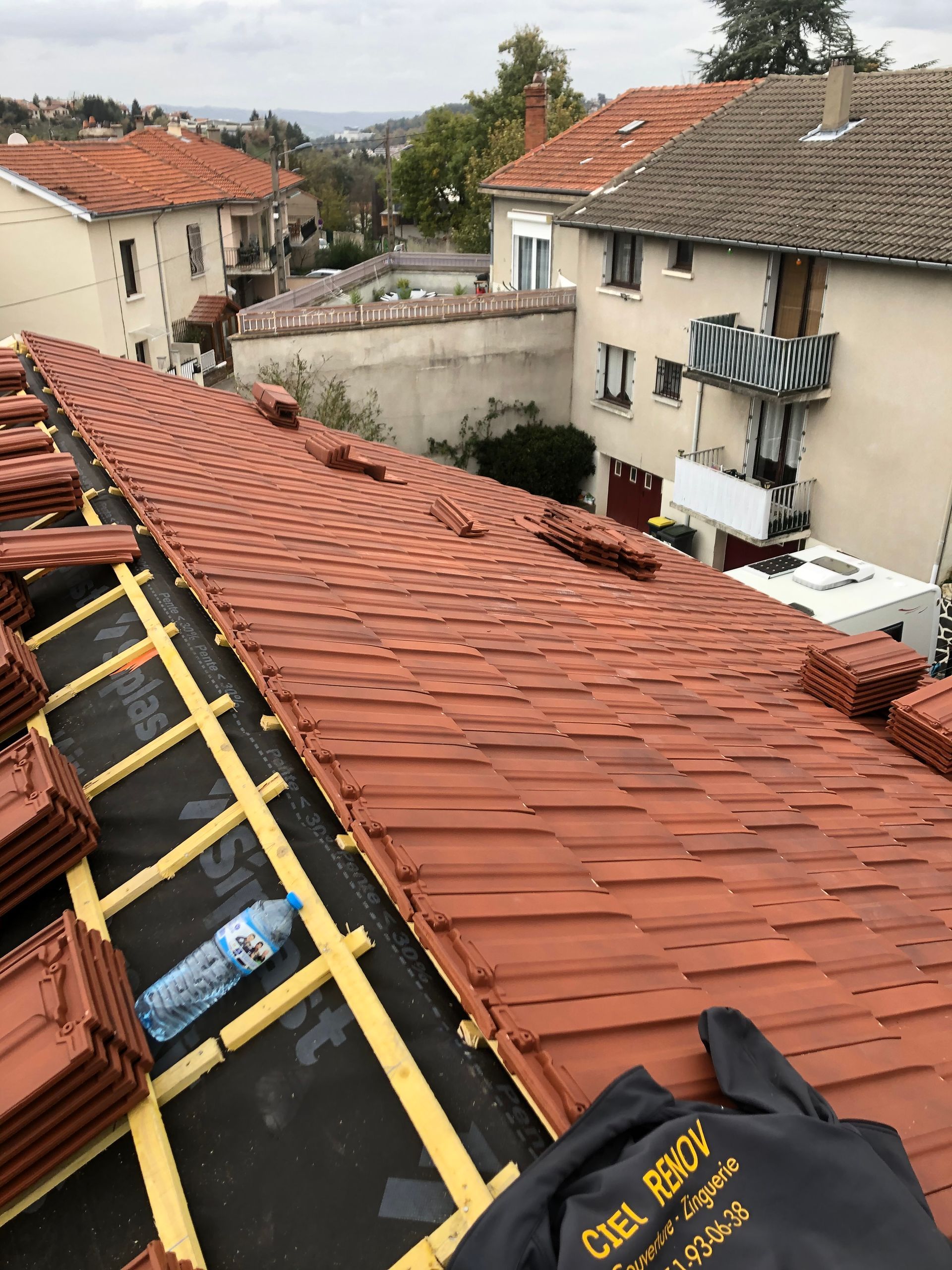 Des couvreurs installent des tuiles rouges sur un toit résidentiel à forte pente, à l'aide de harnais de sécurité.