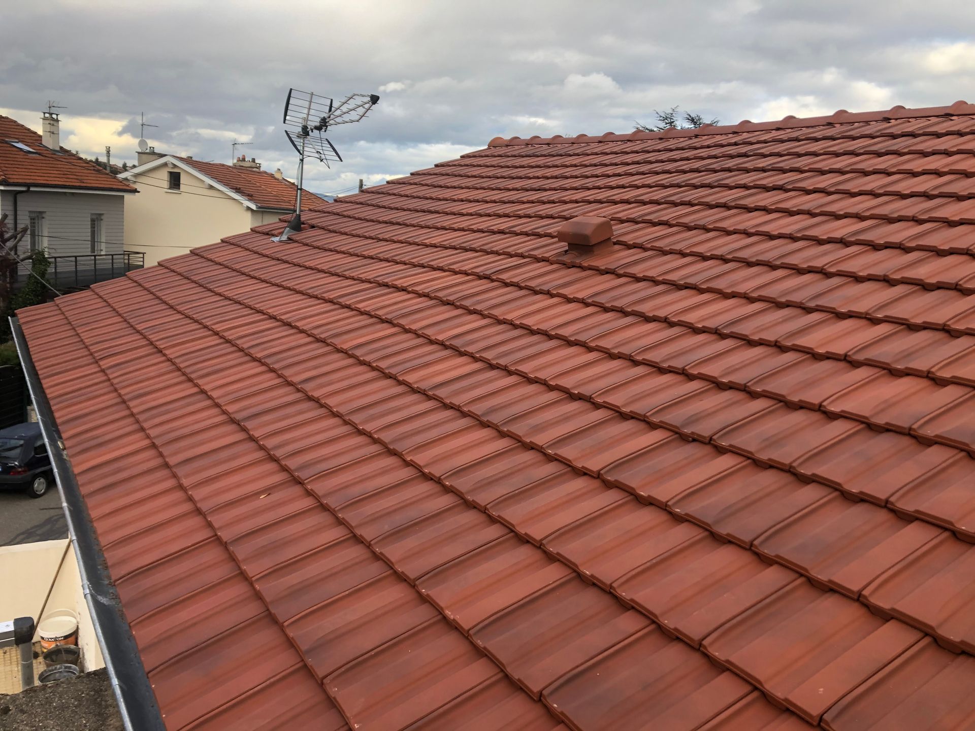 Toit de tuiles en terre cuite rouge sous un ciel nuageux, avec les maisons voisines visibles en arrière-plan.