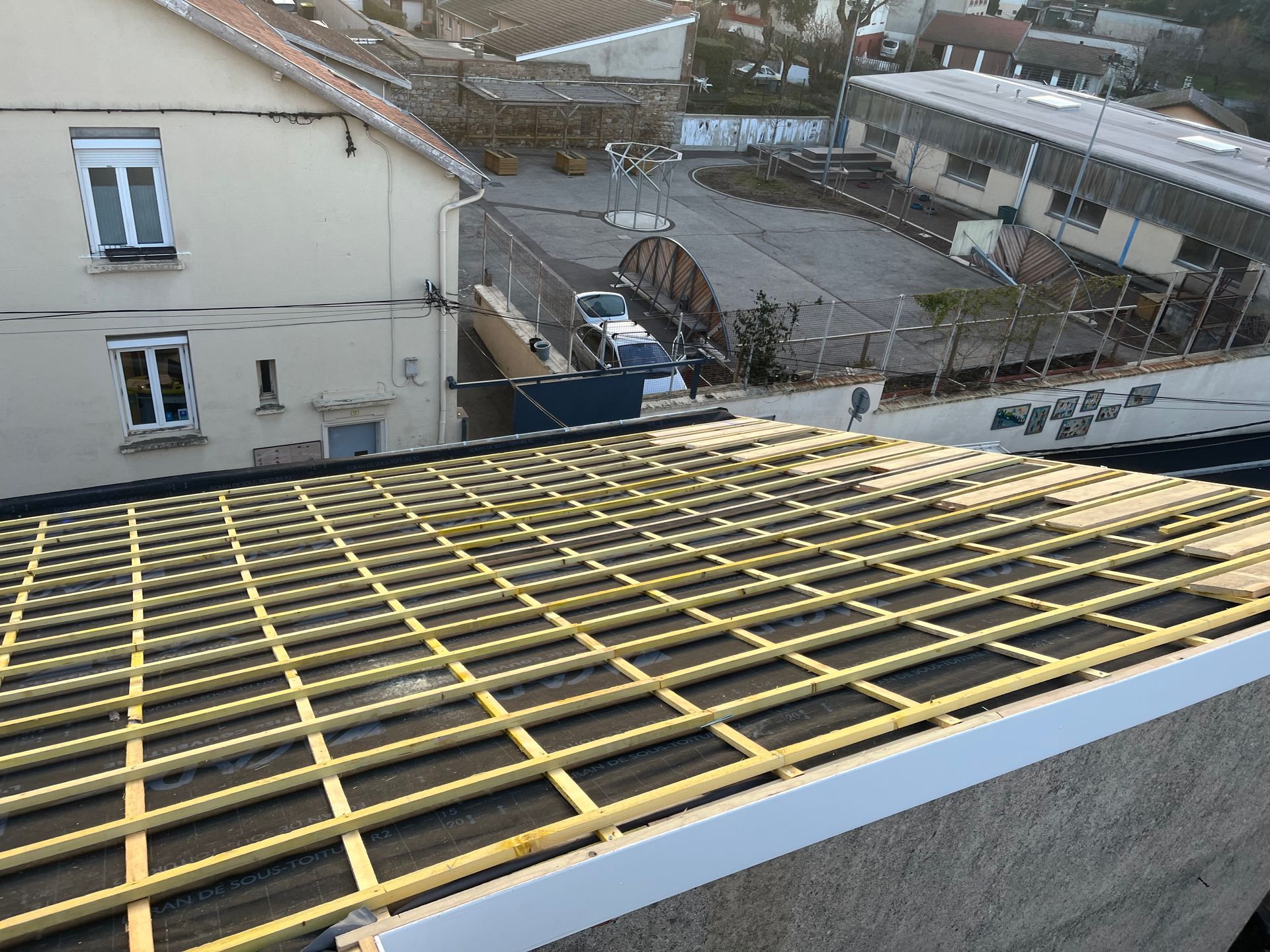 Toiture en construction avec charpente jaune au premier plan et bâtiments à l'arrière-plan.