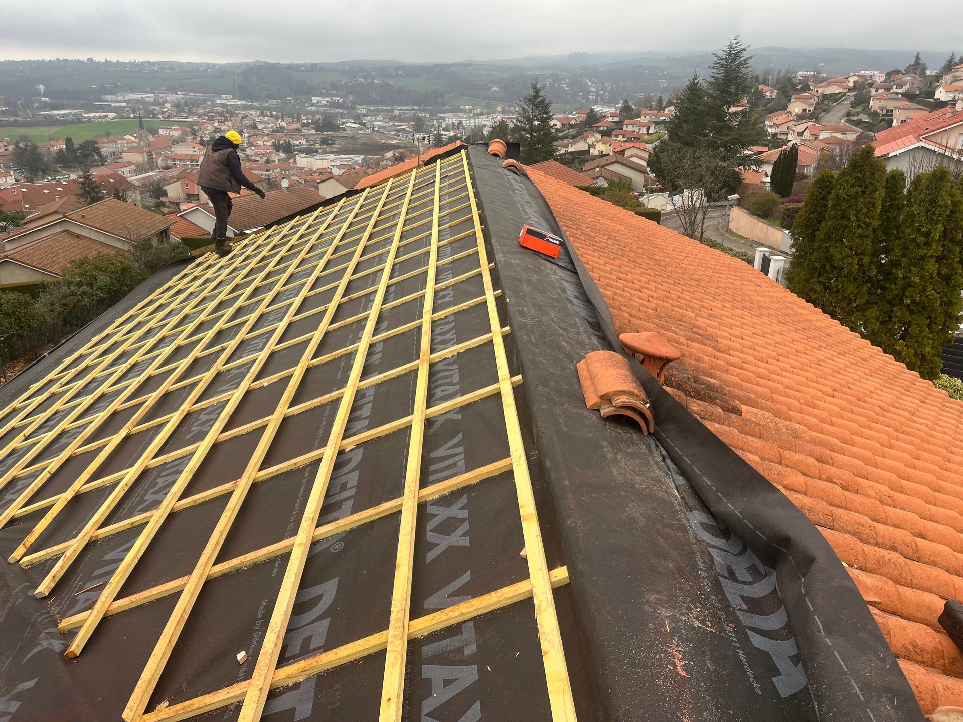Toiture en réparation avec des lattes jaunes et des tuiles orange donnant sur une ville