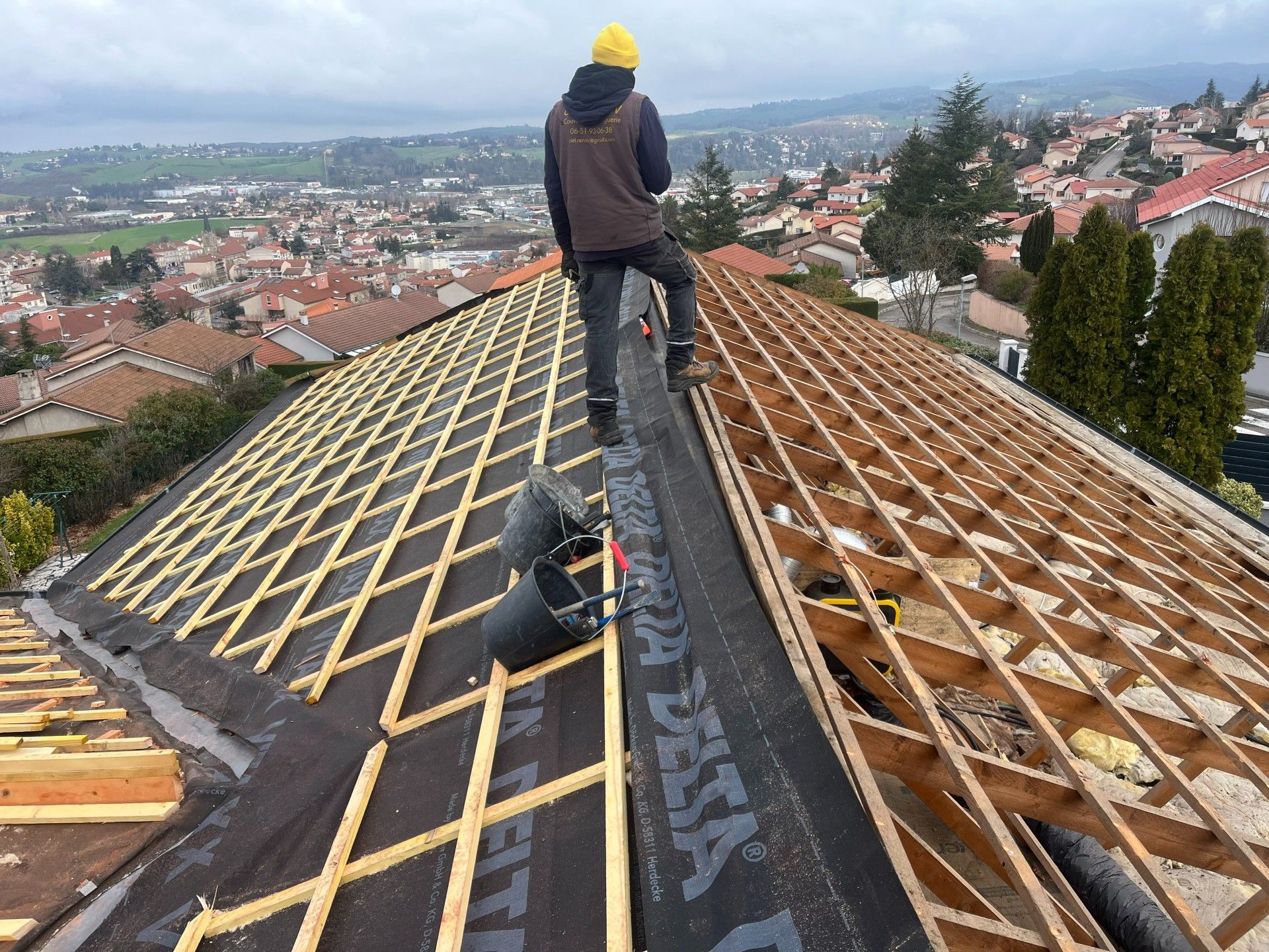 Un couvreur installe une sous-couche sur un toit de tuiles pentu surplombant une ville.