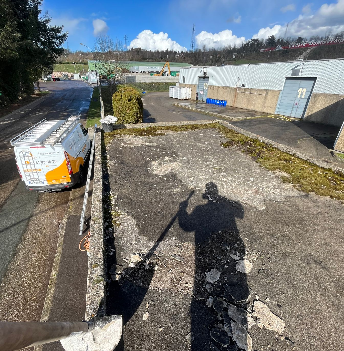 Parking ensoleillé avec une personne dans l'ombre tenant une longue perche à côté d'une zone de gravier et d'une remorque.