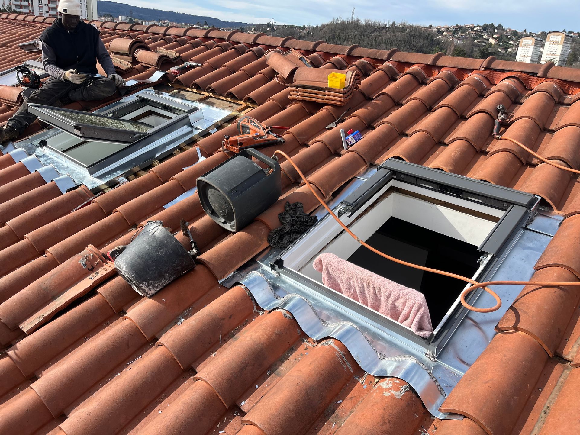 Des couvreurs travaillent sur un toit en tuiles rouges avec des puits de lumière ouverts et des conduits apparents.