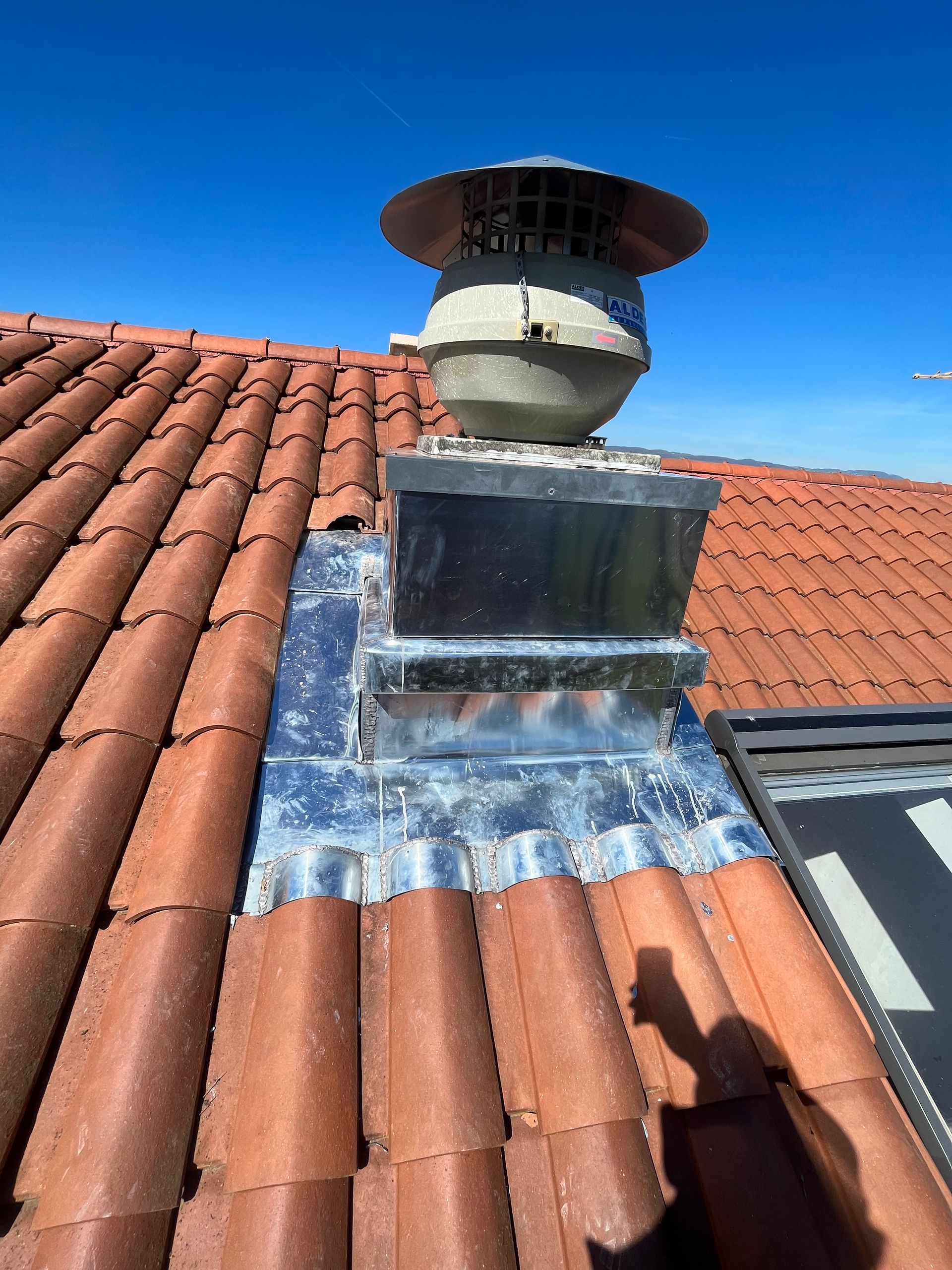 Cheminée de ventilation de toit avec solin métallique sur des tuiles en terre cuite rouge sous un ciel bleu.