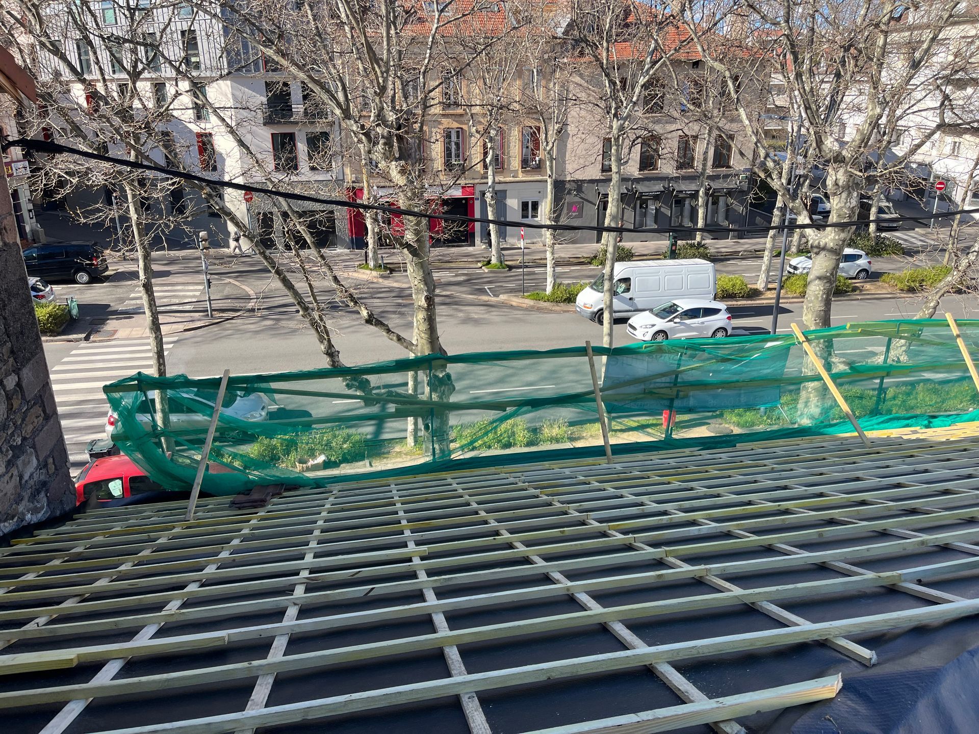 Grille d'échafaudage au premier plan surplombant une rue bordée d'arbres dénudés, d'une camionnette garée et d'immeubles d'habitation.