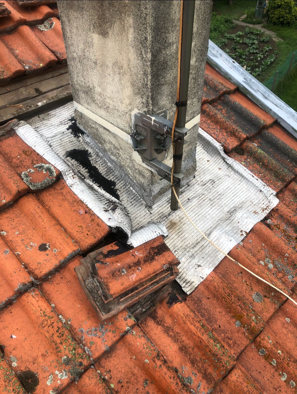 Solin de toiture endommagé autour d'une cheminée sur un toit de tuiles rouges, avec mastic blanc altéré par les intempéries et tuyau métallique apparent.