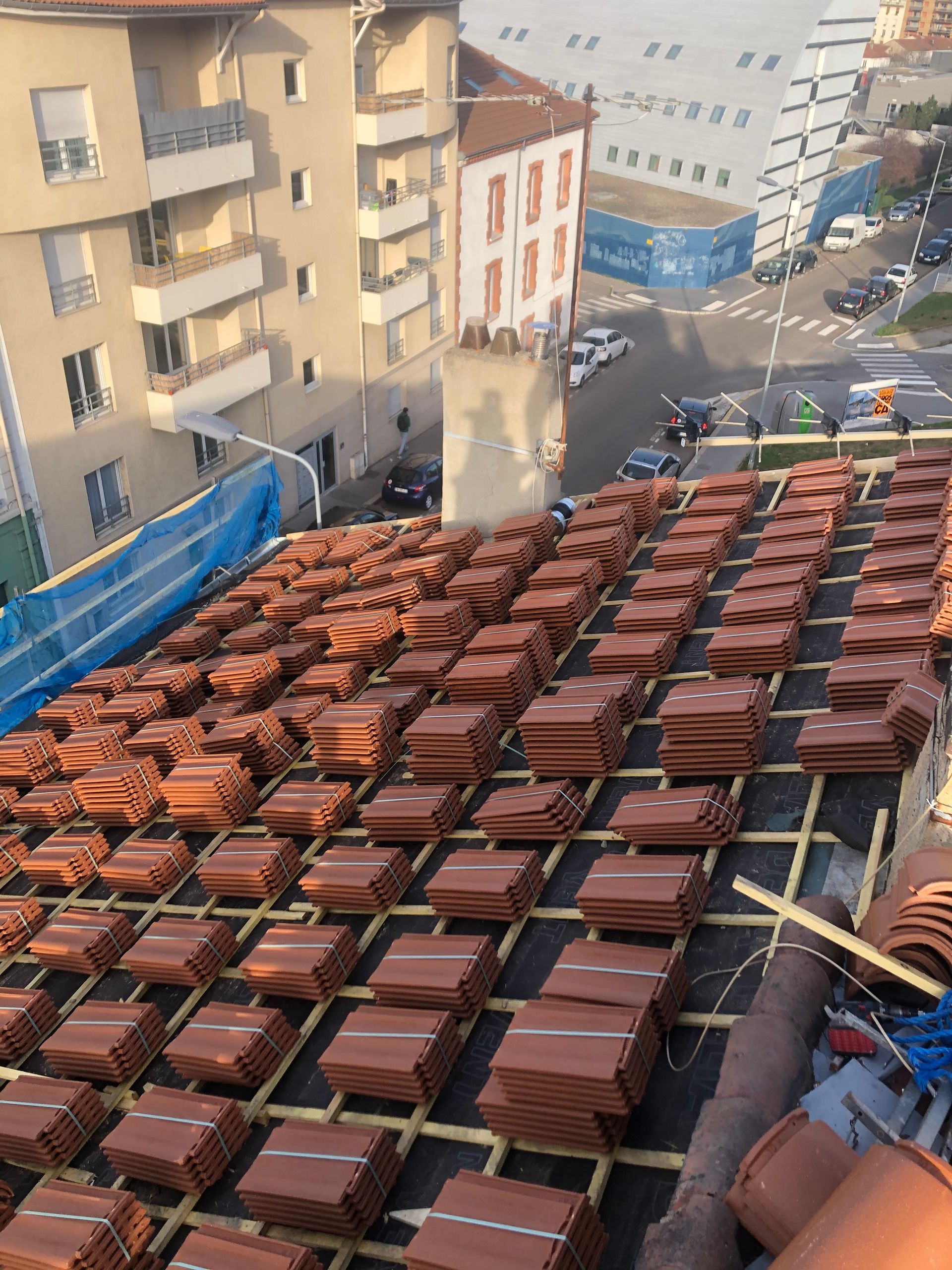 Toit recouvert de rangées de briques en terre cuite, jouxtant des immeubles d'habitation et des filets de sécurité bleus.