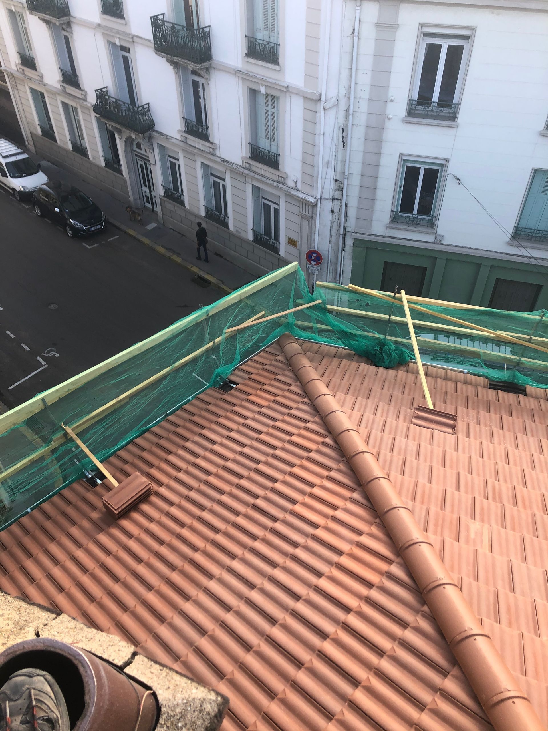 Toit-terrasse recouvert de tuiles en terre cuite rouge et de filets de sécurité verts donnant sur une rue étroite et des bâtiments blancs.