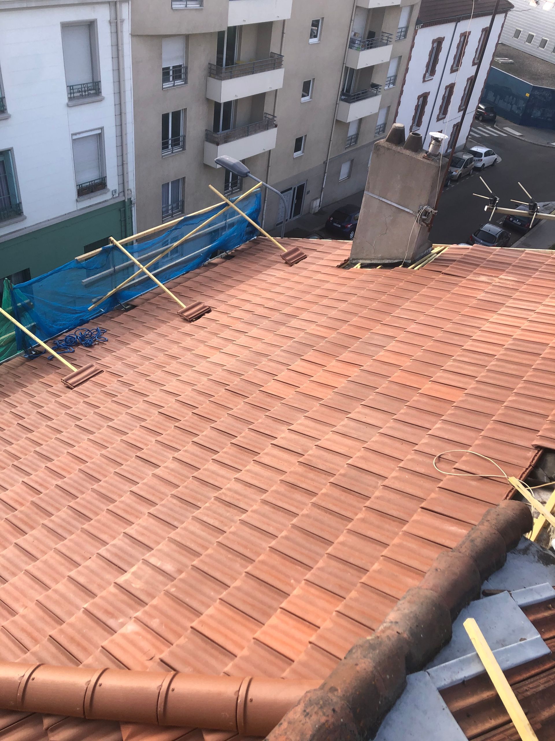 Toit-terrasse avec toit de tuile rouge, bâche bleue et immeubles d'appartements à proximité, en milieu urbain.