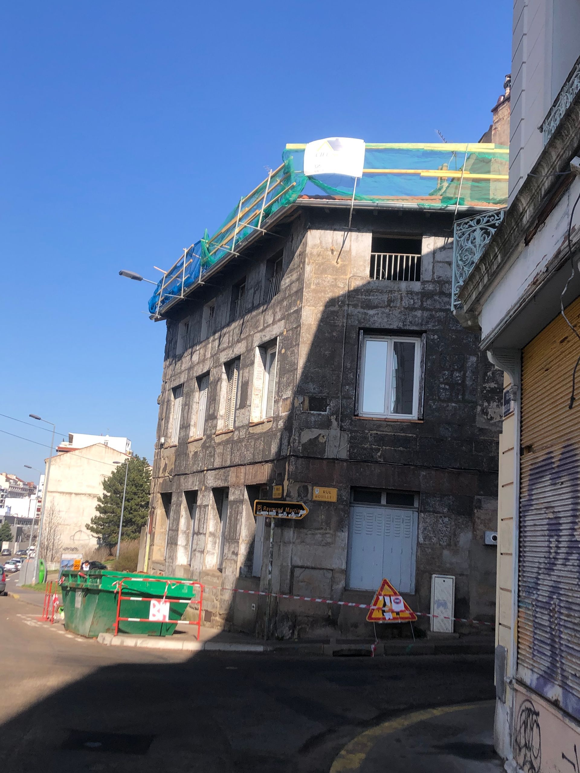 Vieux bâtiment en pierre aux fenêtres condamnées, toit en échafaudages recouvert de végétation et une personne vêtue d'orange près de l'entrée.