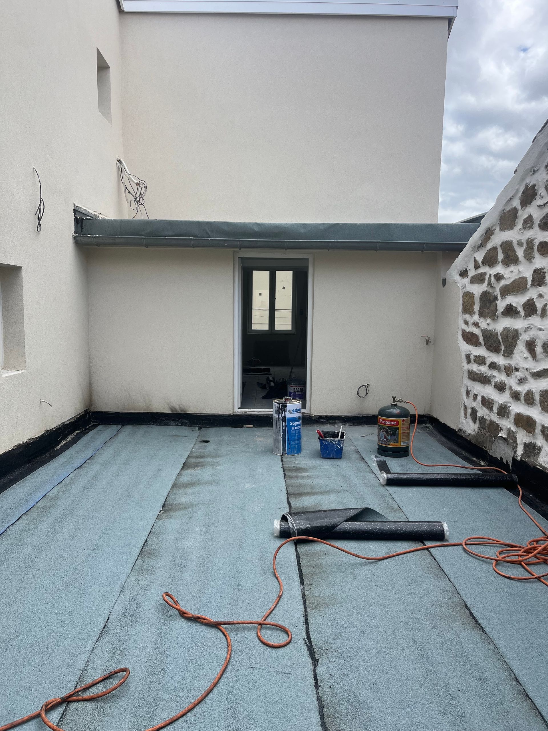 Toit plat en rénovation avec membrane d'étanchéité bleue, outils et porte noire sur un mur blanc.