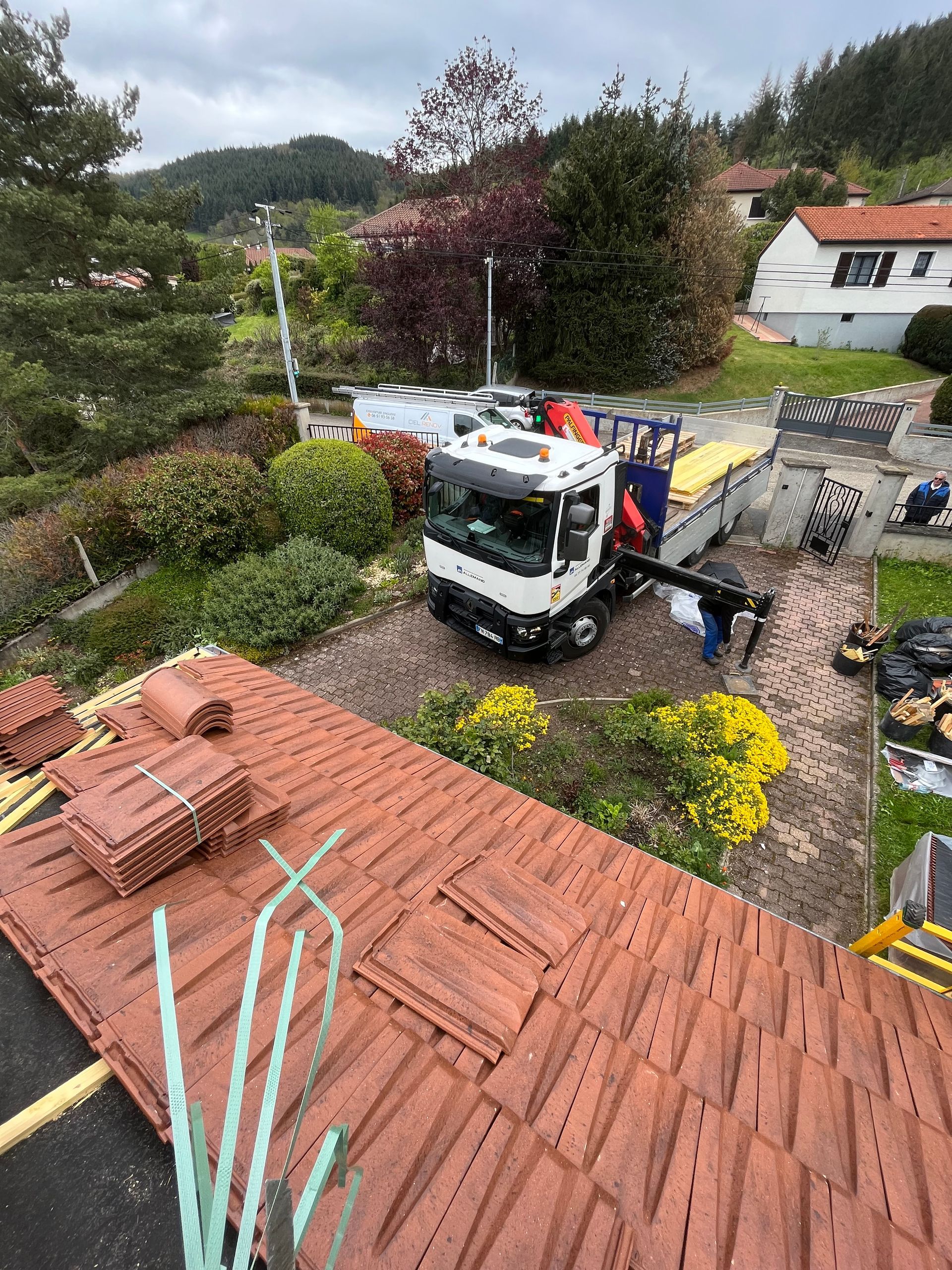 Un ouvrier décharge des matériaux sur une grue mobile dans la cour d'une maison aux toits de tuiles rouges et aux fleurs.