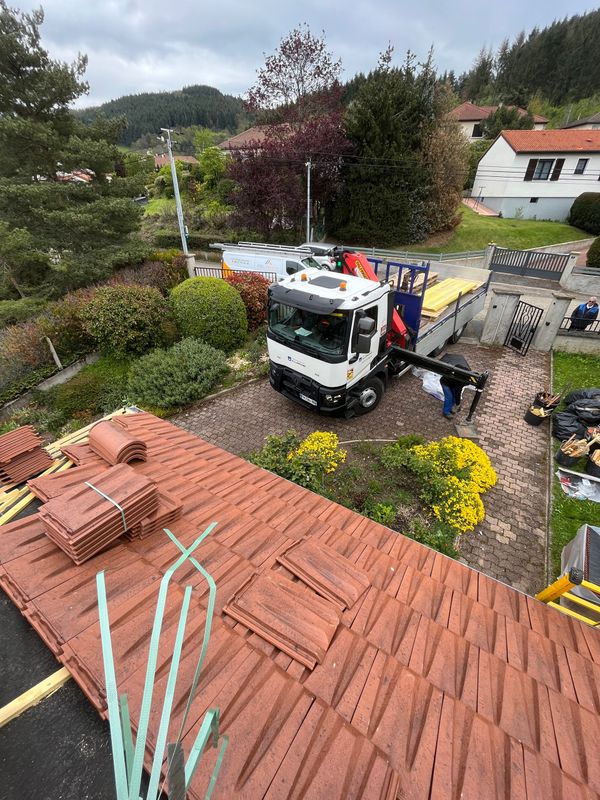 Un ouvrier décharge des matériaux sur une grue mobile dans la cour d'une maison aux toits de tuiles rouges et aux fleurs.