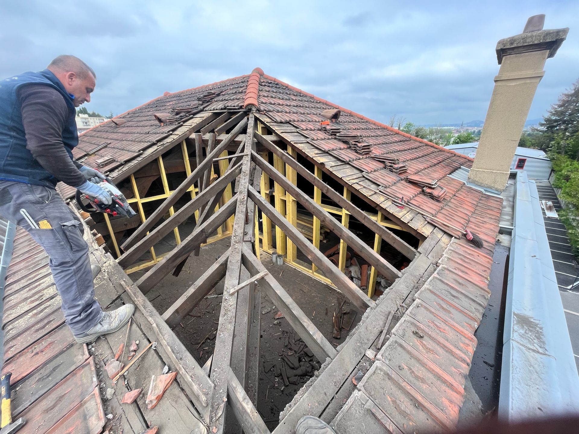 Un ouvrier répare un toit résidentiel endommagé, avec des chevrons en bois apparents et des tuiles en terre cuite rouge, sous un ciel couvert.