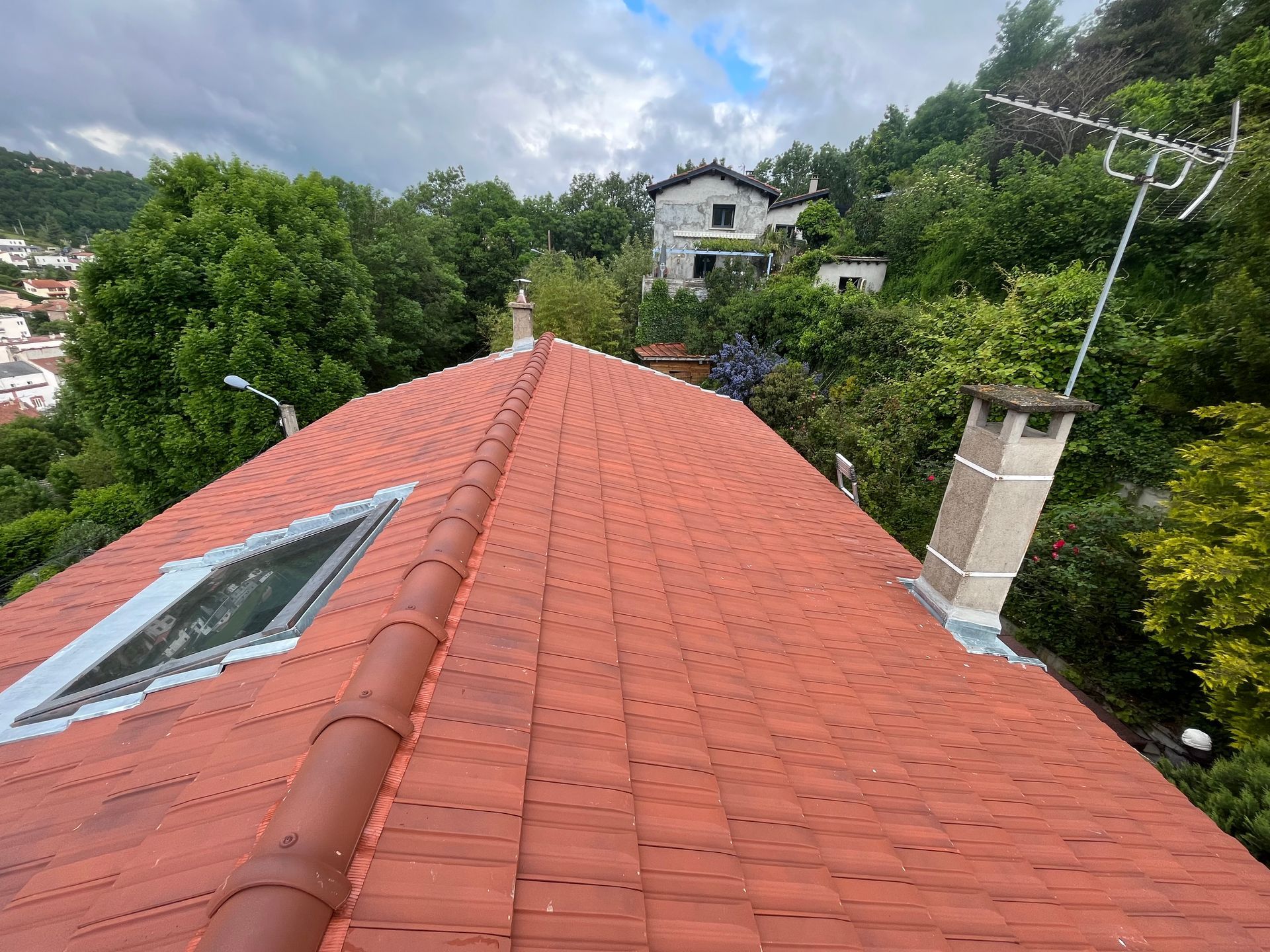Toit de tuiles rouges donnant sur des collines verdoyantes et des maisons sous un ciel nuageux.