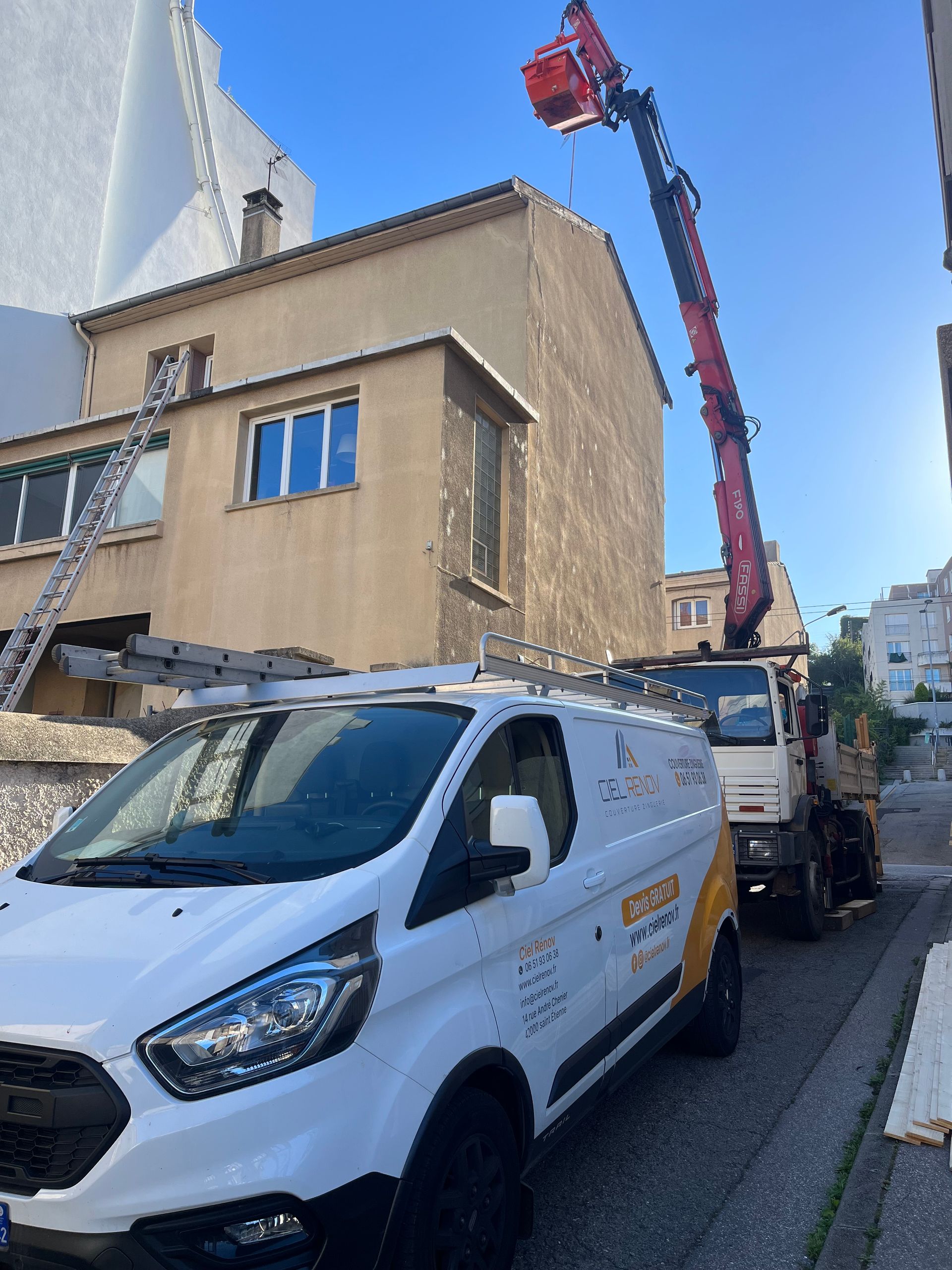 Camionnette blanche et nacelle élévatrice devant un bâtiment dans une rue étroite sous un ciel bleu.