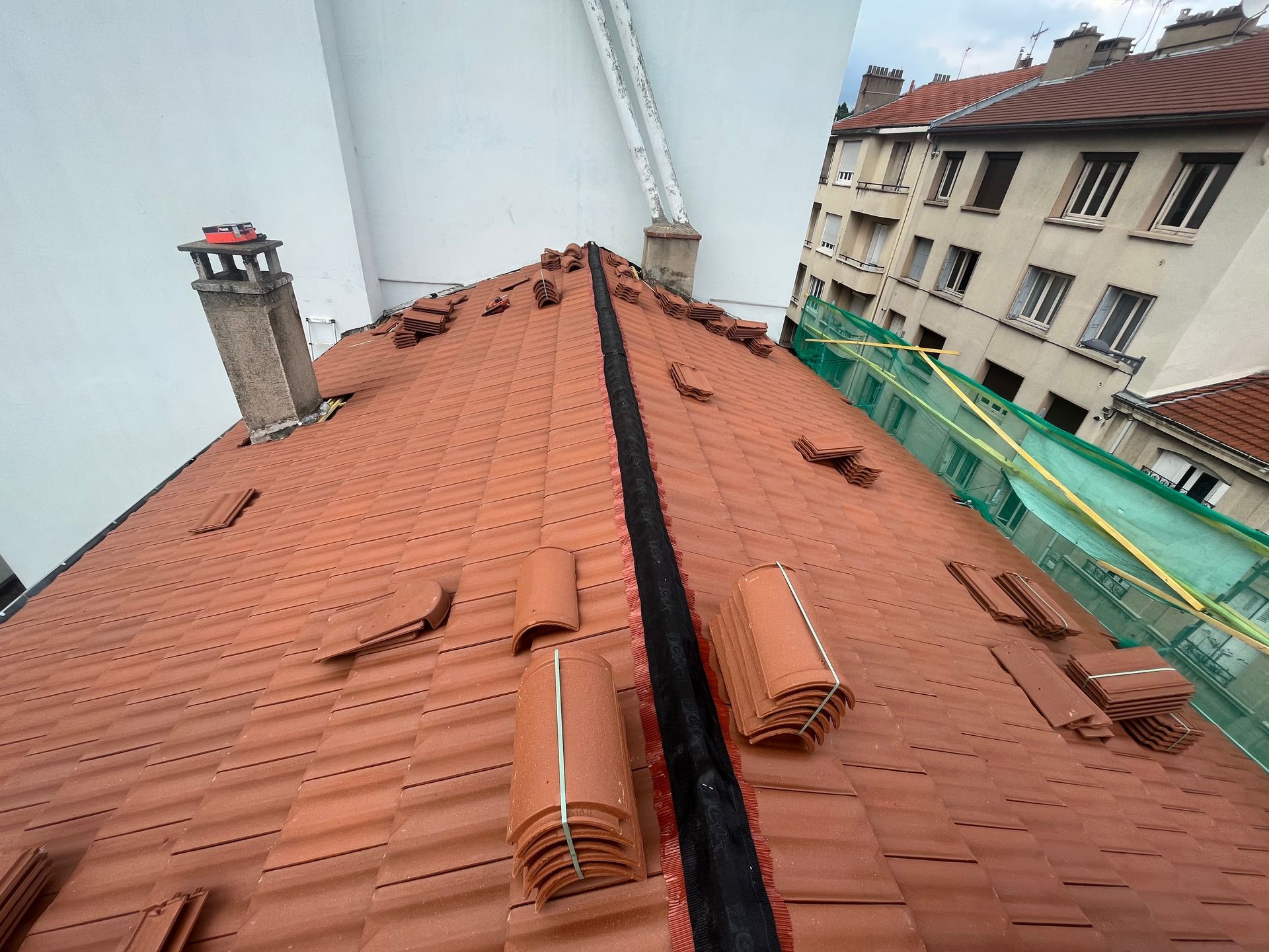 Toit en tuile rouge avec une arête centrale, cheminée et échafaudages à côté des bâtiments voisins.