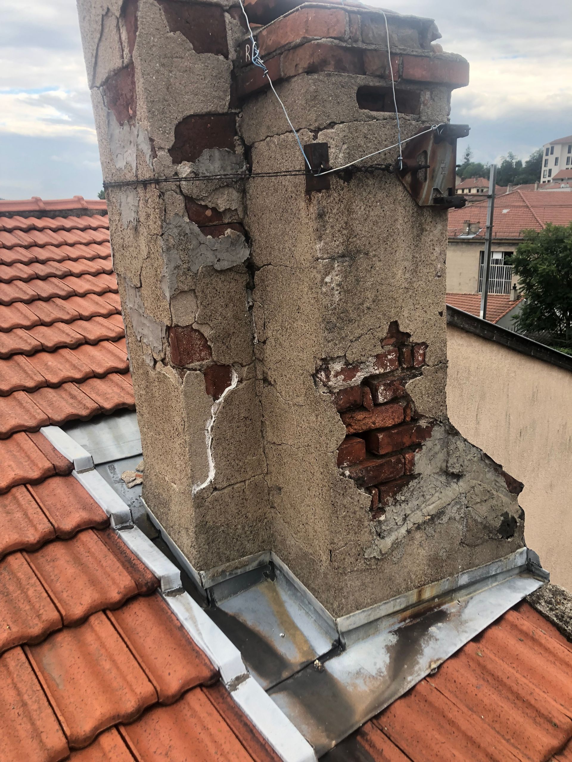 Cheminée en brique endommagée sur un toit de tuile rouge, avec plâtre et solins métalliques effrités à la base