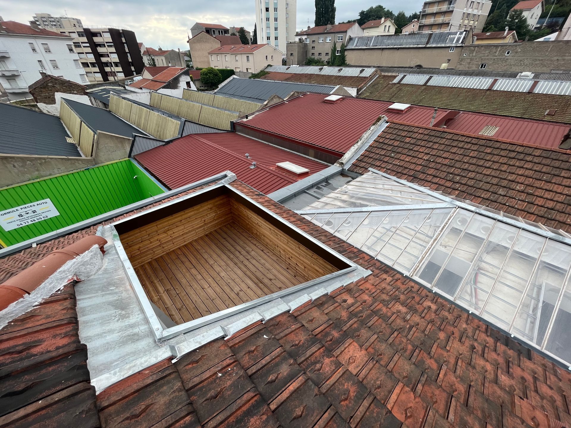 Vue depuis le toit avec des toitures en tuiles rouges et grises, une ouverture terrasse tropézienne en bois et la silhouette de la ville en arrière-plan.
