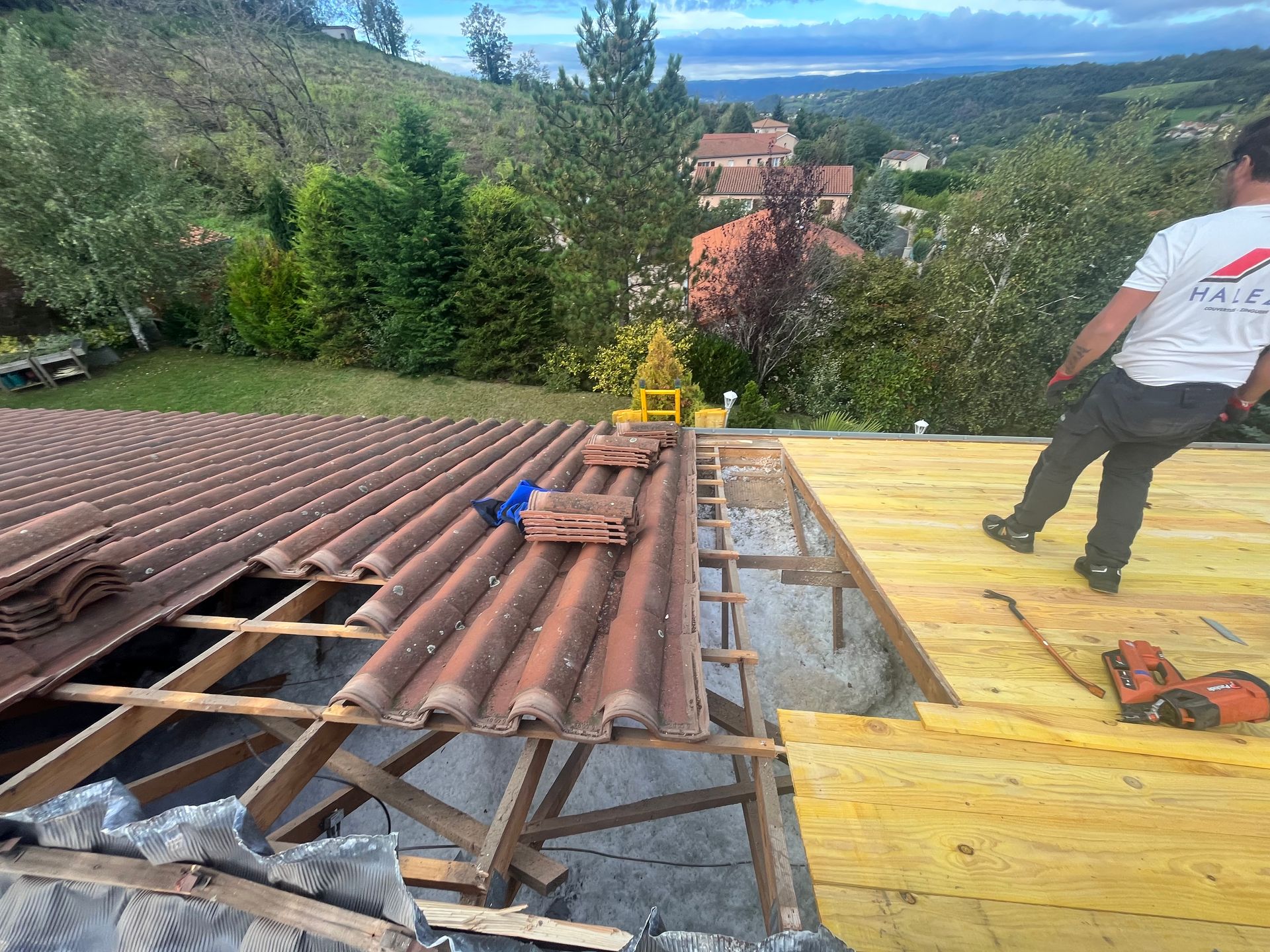 Des couvreurs remplacent un toit de tuiles brunes à côté d'une sous-couche jaune, avec vue sur la montagne au loin.