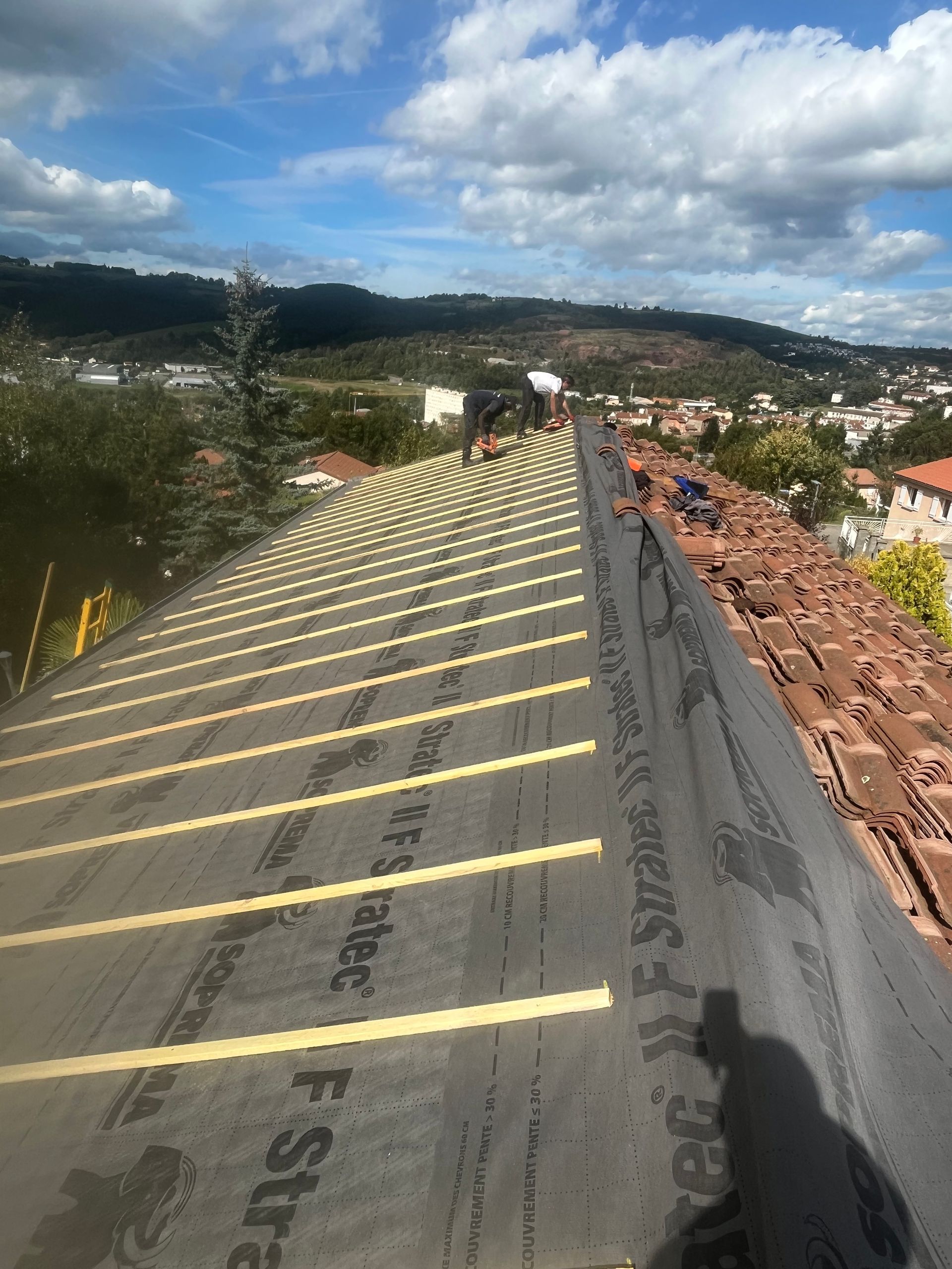 Toit en construction avec des lignes de sécurité jaunes, toits de la ville et montagnes sous un ciel nuageux.