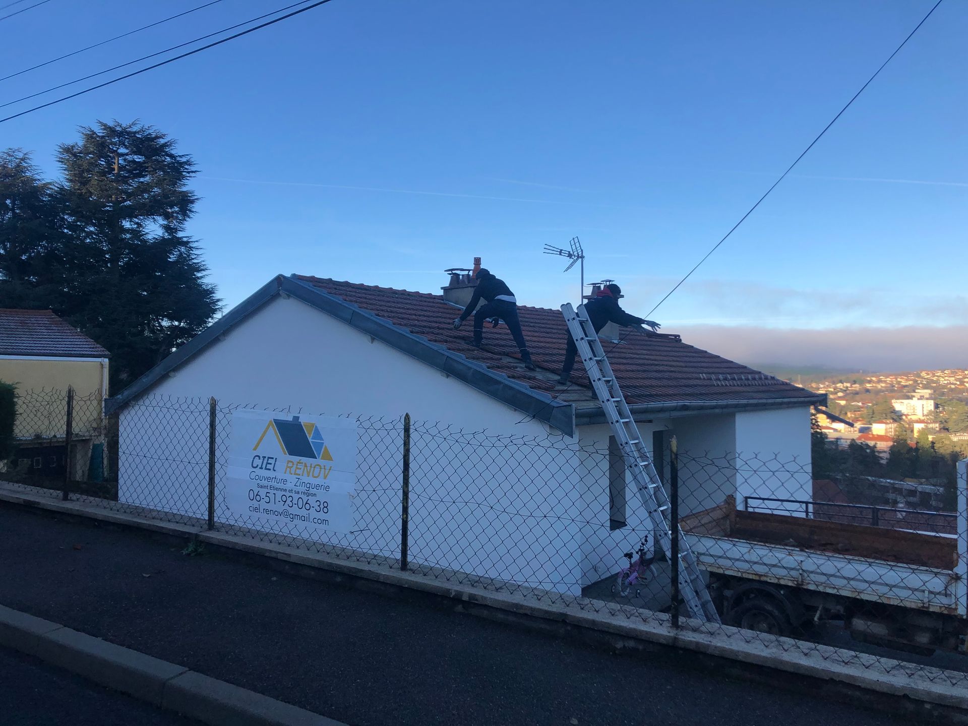 Deux ouvriers sur un toit de tuiles à côté d'un bâtiment blanc au crépuscule, avec une échelle et une vue sur la colline.
