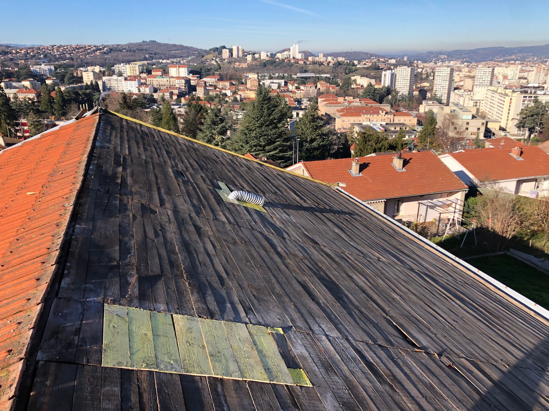 Toiture recouverte de bardeaux sombres et patinés et de tuiles rouges, surplombant un paysage urbain dense sous un ciel dégagé.