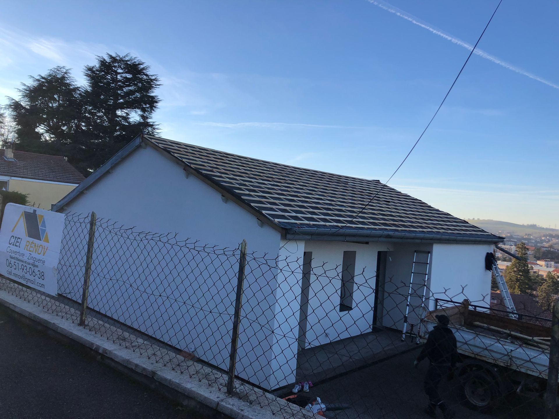 Maison blanche au toit de tuiles à flanc de colline, cour clôturée, et une personne près de l'entrée au coucher du soleil.