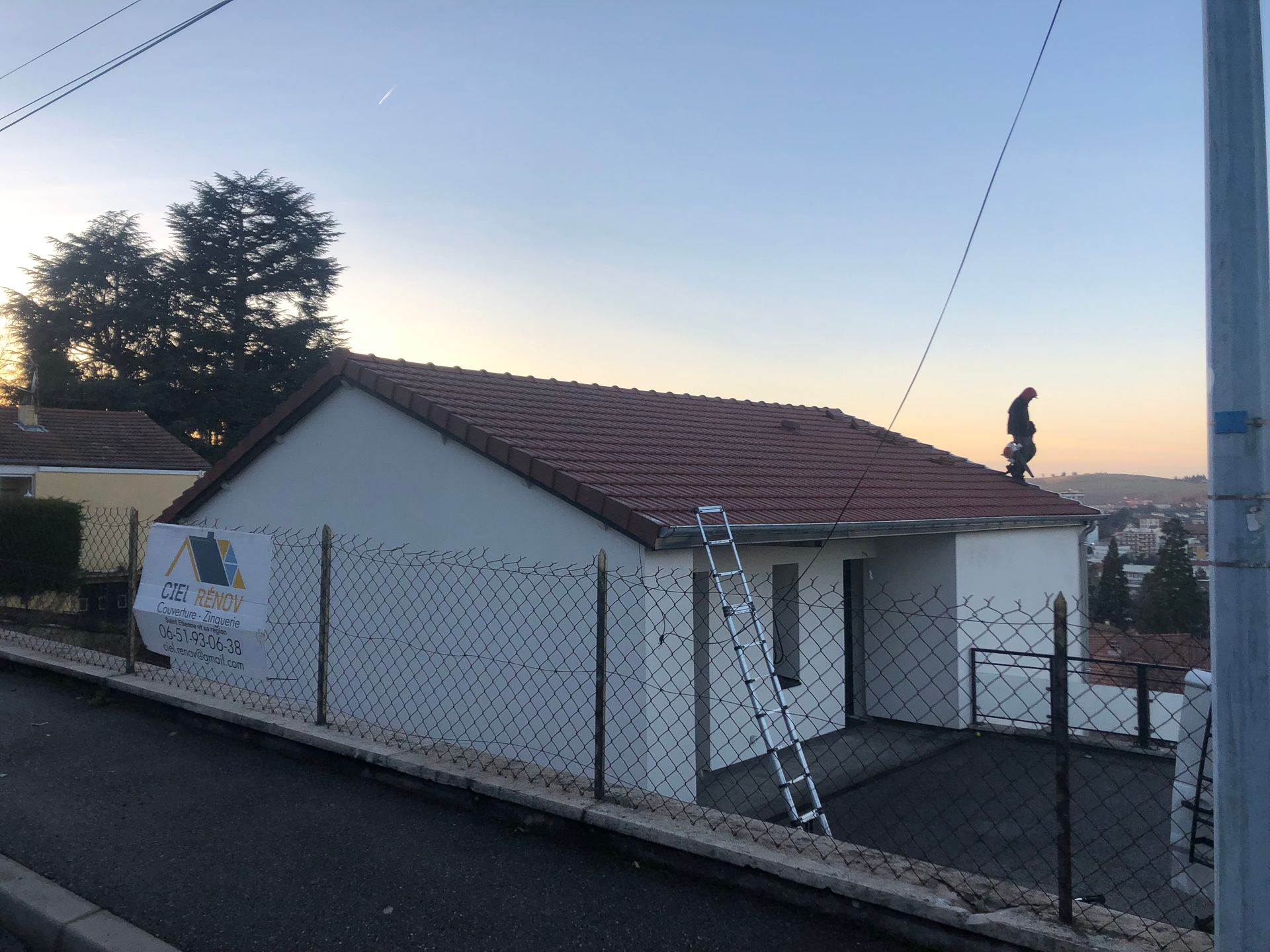 Petite maison blanche au toit rouge derrière une clôture en grillage au coucher du soleil, vue depuis une rue en pente.