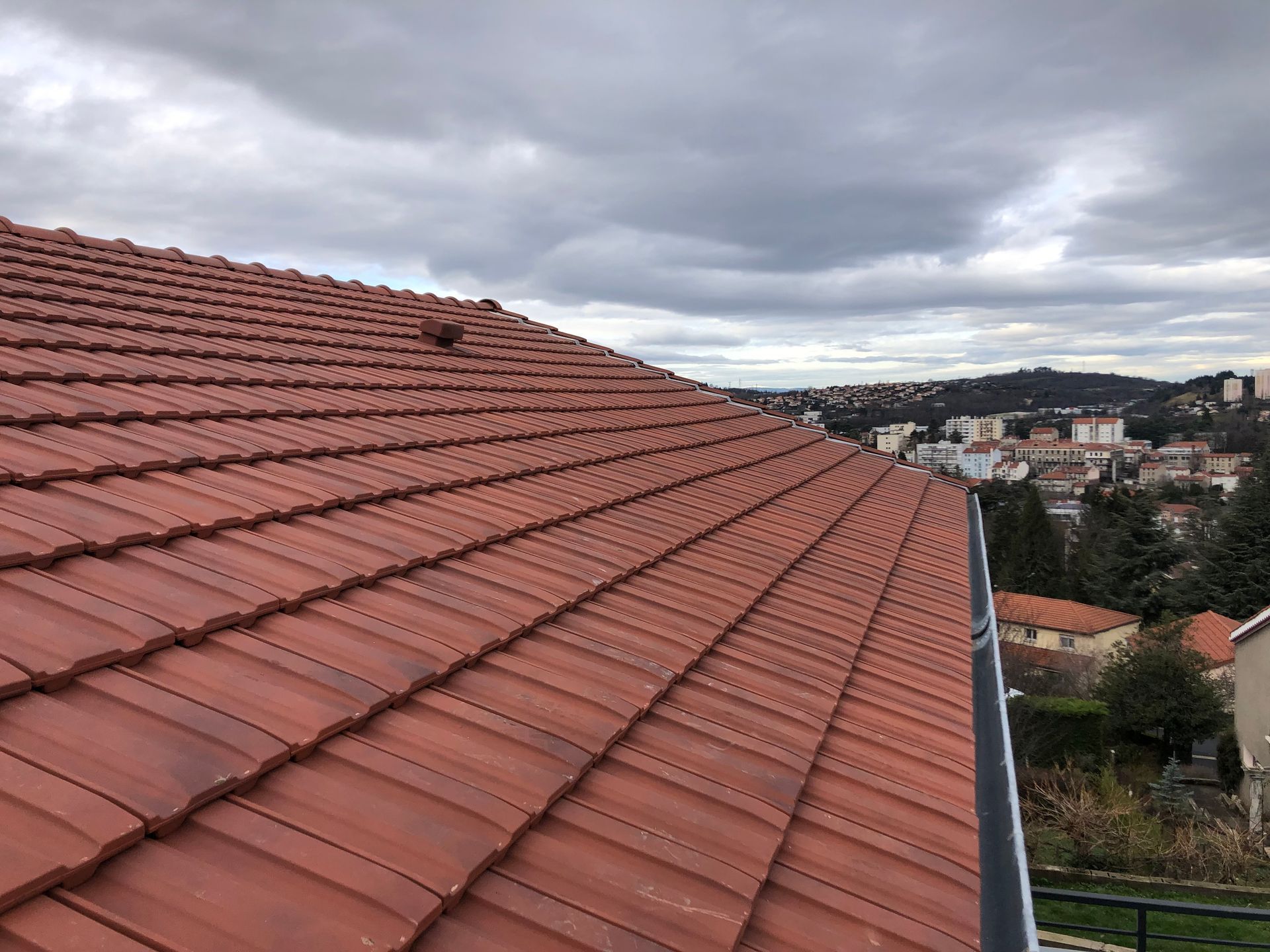 Toit de tuile rouge donnant sur un paysage urbain nuageux et des collines au loin.