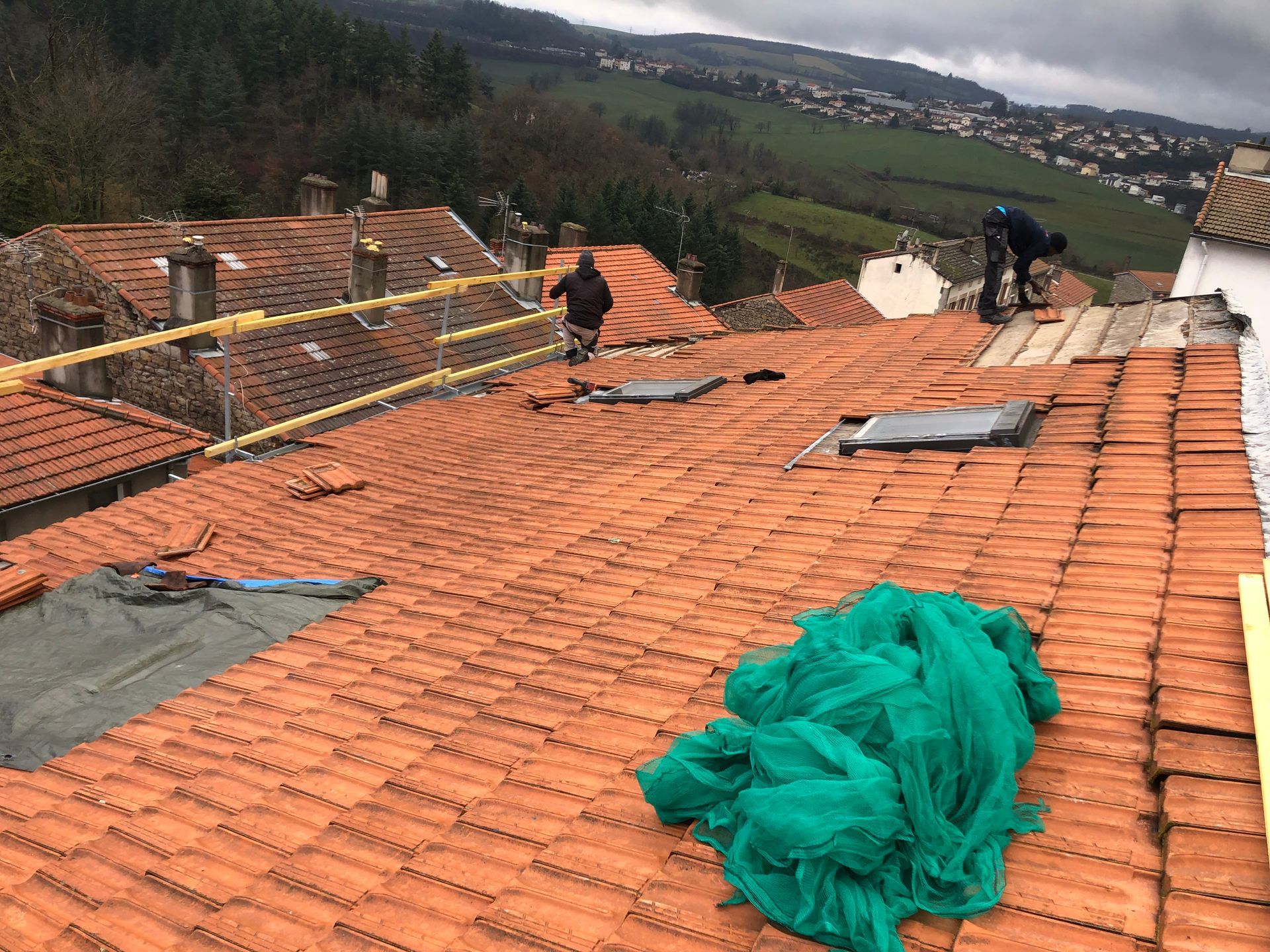 Des ouvriers réparent un toit de tuiles en terre cuite à l'aide de filets de sécurité, surplombant une vallée rurale.