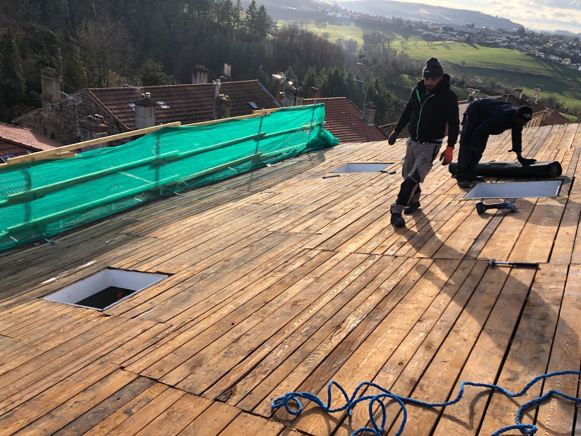 Des ouvriers installent un platelage de toit en bois sur un toit en pente, avec des filets de sécurité verts et une vue sur la vallée.
