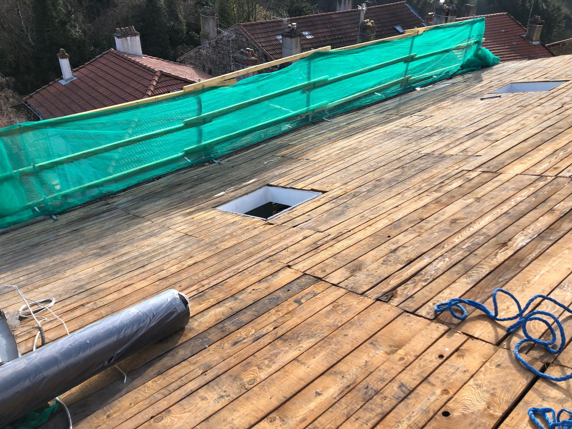 Toiture en réparation avec des planches, filet de sécurité vert, ouvertures de puits de lumière et un tuyau bleu enroulé.