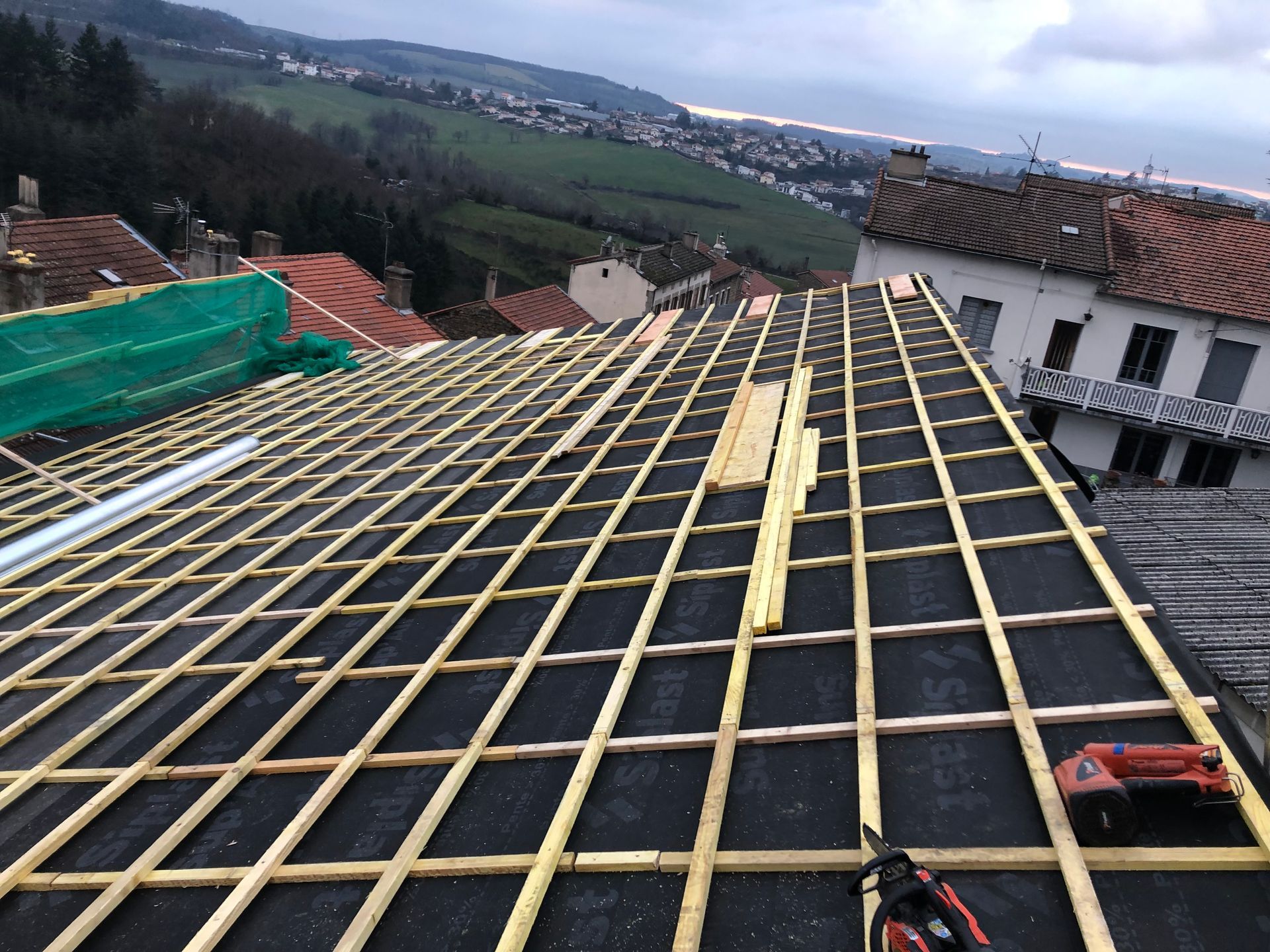 Toiture en construction avec liteaux en bois et sous-couche noire, donnant sur un quartier rural à flanc de colline.
