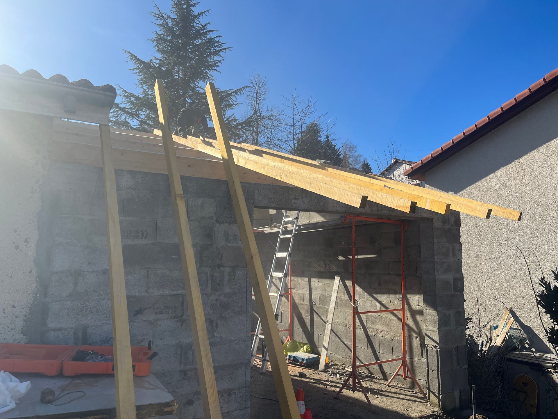Charpente de toit en bois en cours de construction sur une structure murale en blocs de béton, à l'extérieur par une journée ensoleillée.