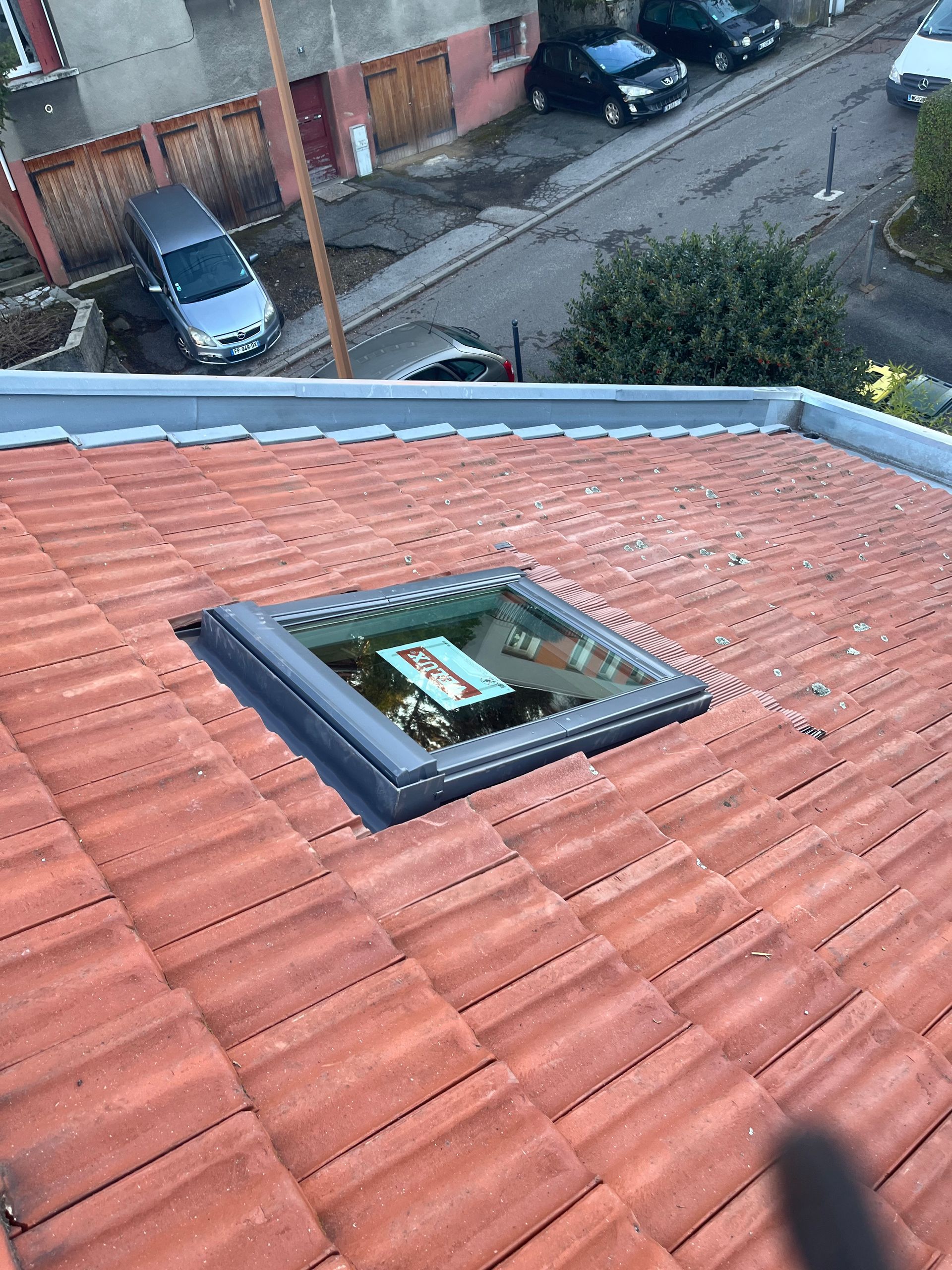 Toit-terrasse avec tuiles rouges et puits de lumière donnant sur une rue résidentielle et des voitures garées.