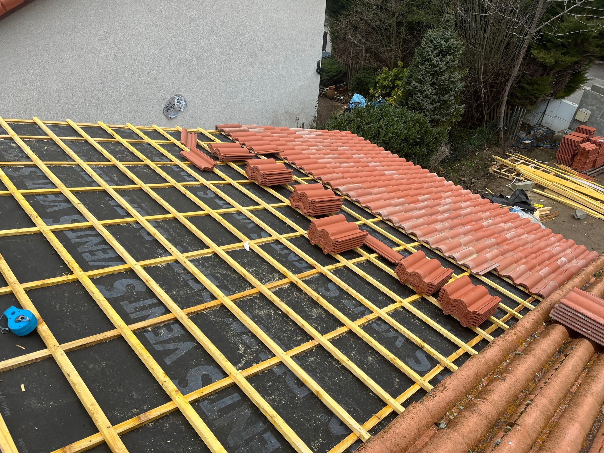 Toiture en construction avec des liteaux jaunes et des piles de tuiles rouges sur une toiture partiellement couverte.