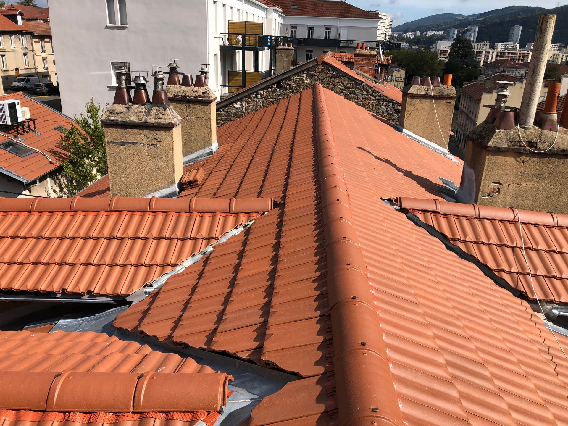 Vue en plongée d'un toit de tuile en terre cuite rouge avec plusieurs cheminées, se détachant sur un fond de bâtiments urbains.