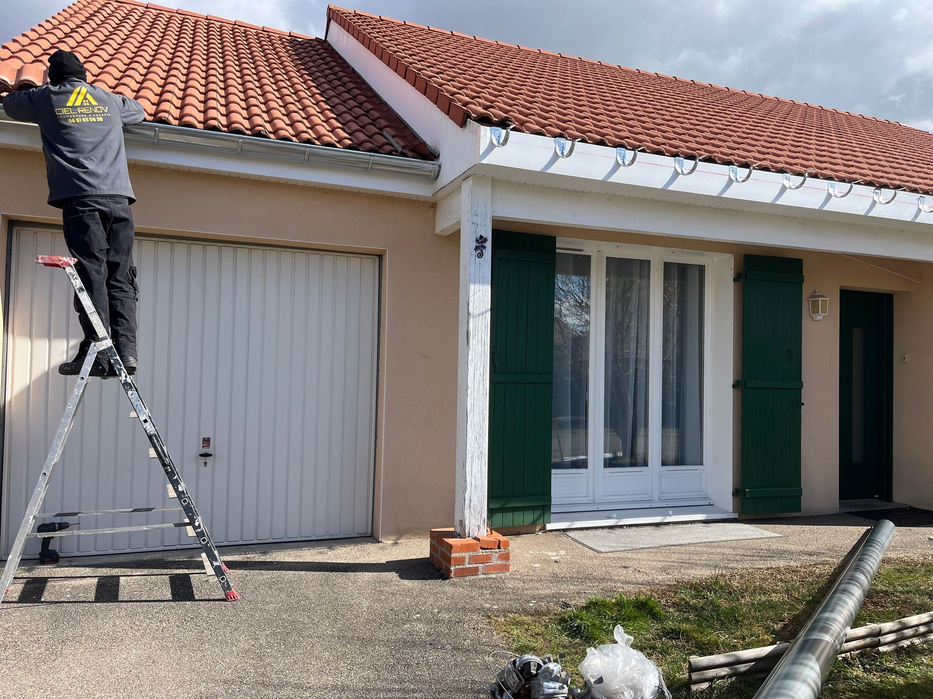 Un ouvrier, juché sur une échelle, répare le toit de tuiles rouges d'une maison blanche aux volets verts.