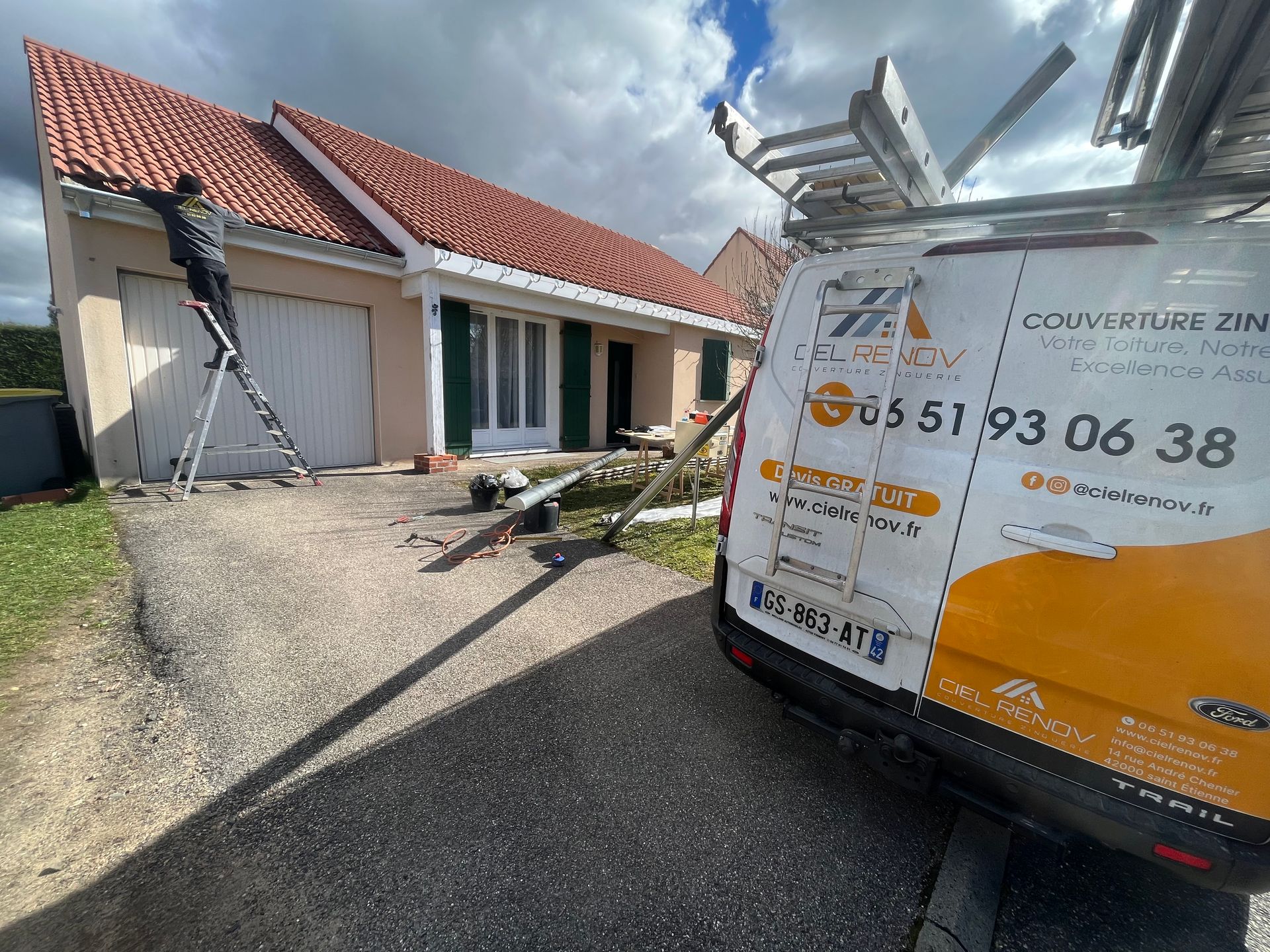Un ouvrier sur l'échelle, répare le toit d'une maison en tuiles rouges à côté d'une camionnette de service blanche garée dans une allée.